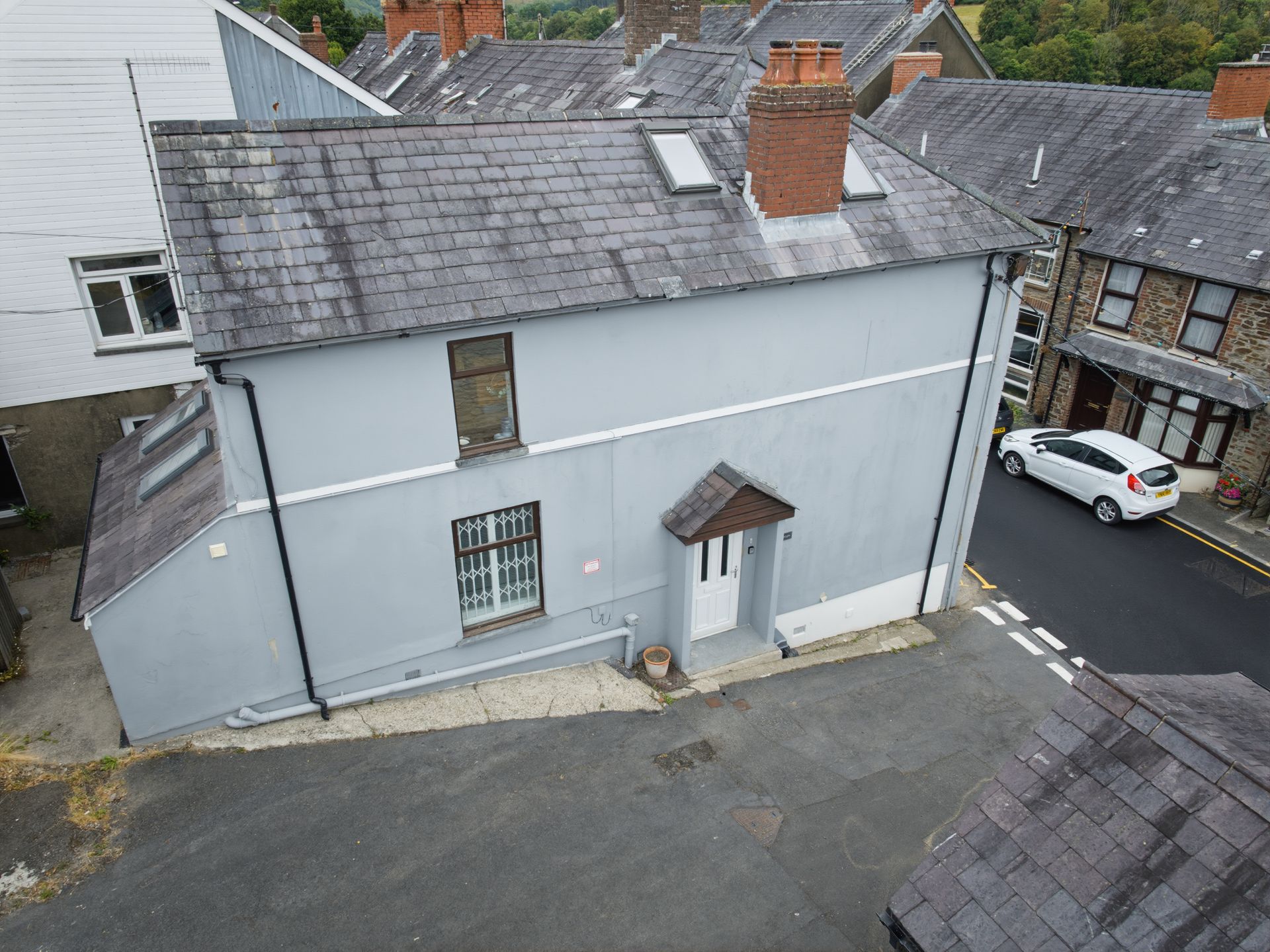 Aerial view of a gray stone house with a slate roof and parked white car on a narrow street.
