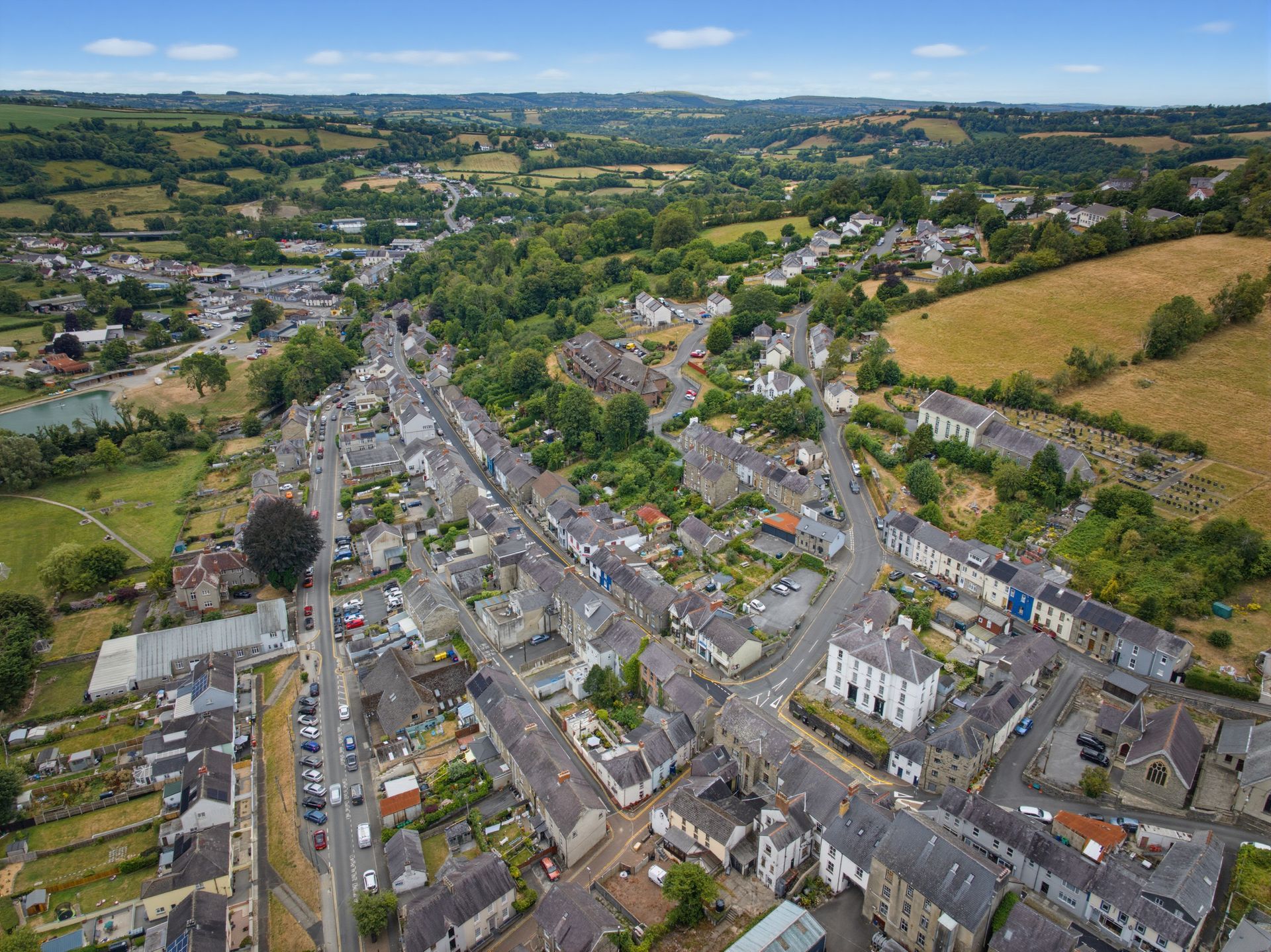 Aerial view of a village road lined with houses, fields, and green hills under a clear sky