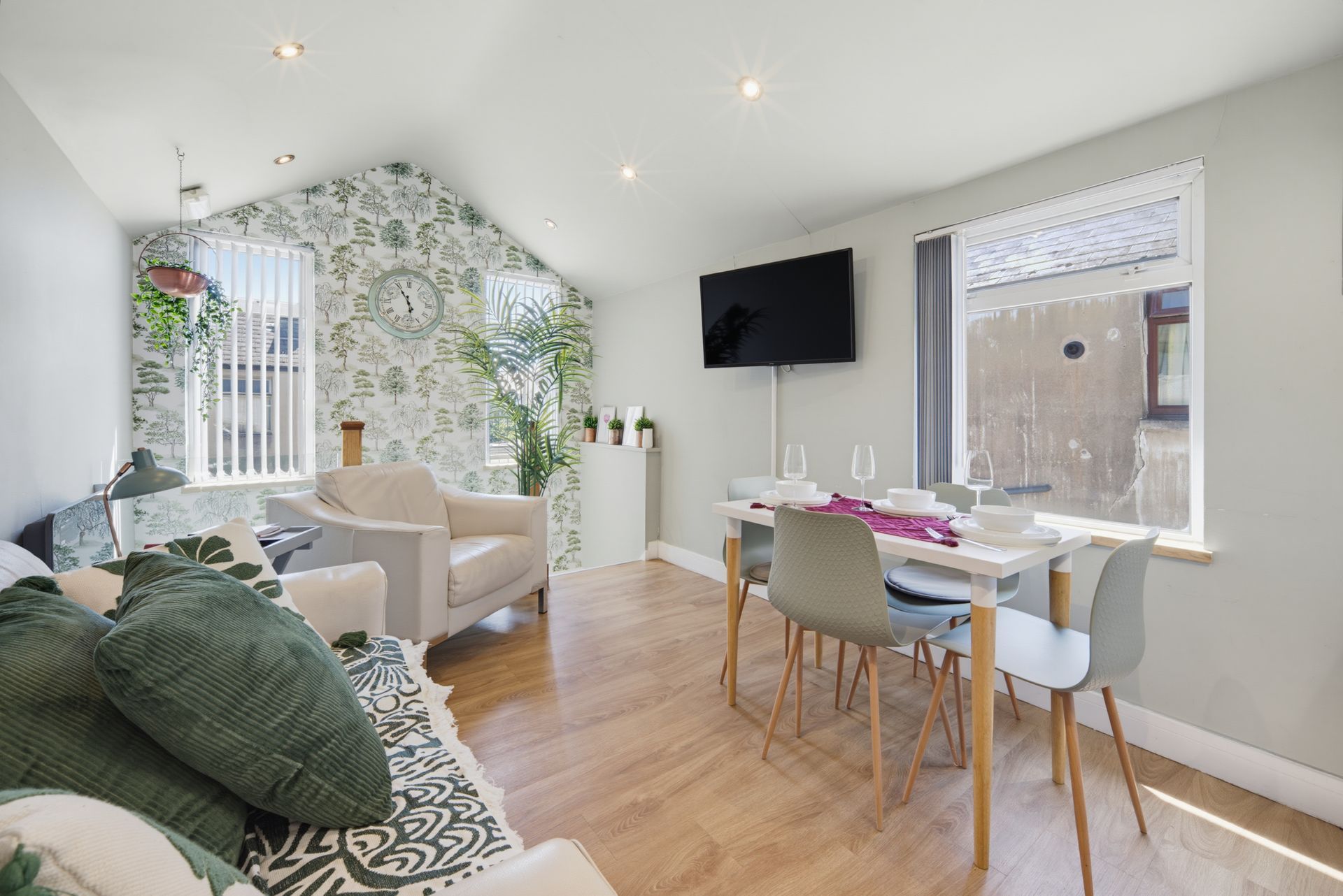 Bright living-dining room with patterned sofa, dining table, wall TV, and floral accent wall under skylights