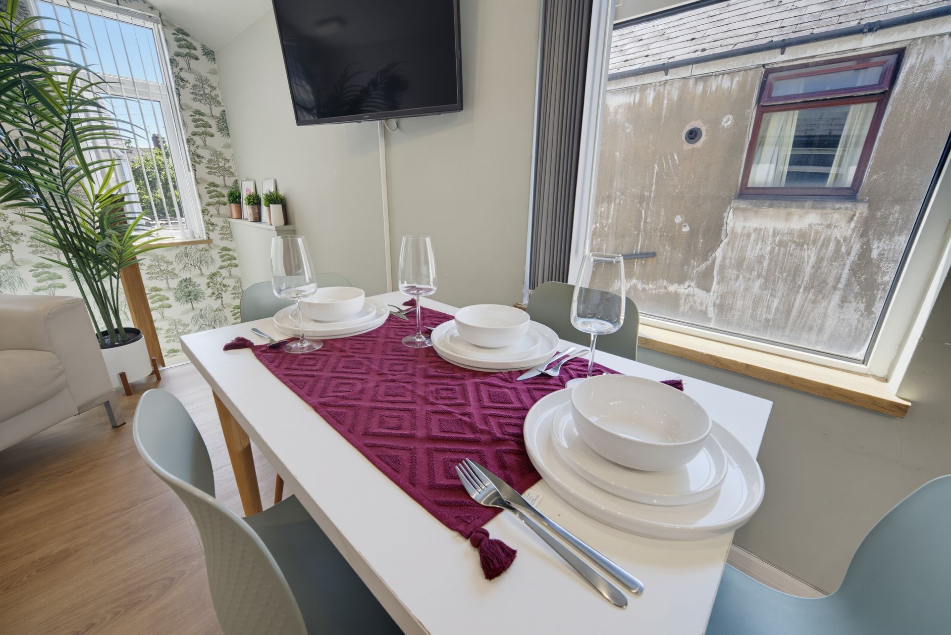 Dining table set with white plates, glasses, and a burgundy runner beside a sunlit window