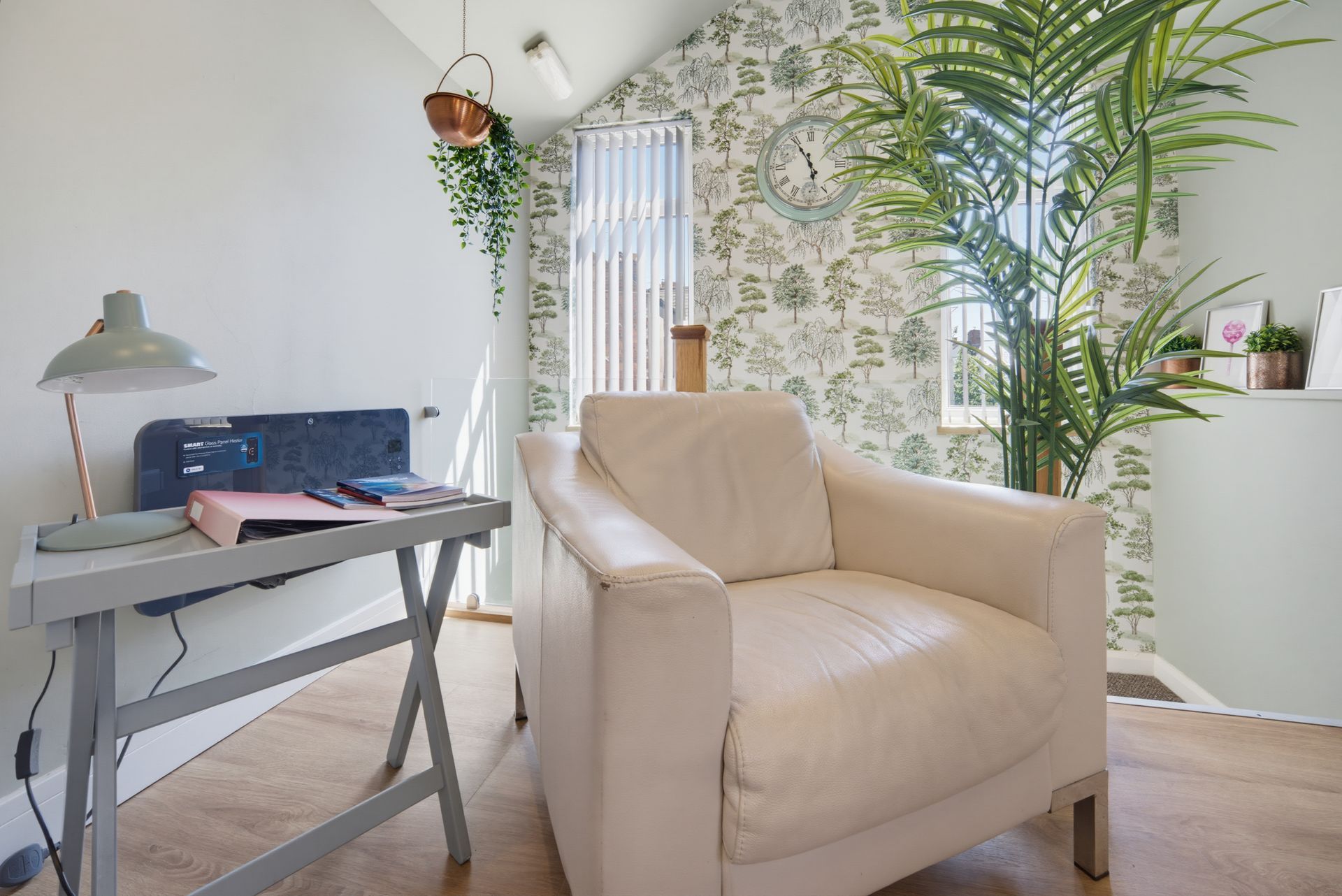 Bright home office with beige armchair, desk, laptop, lamp, and lush green plants by a window