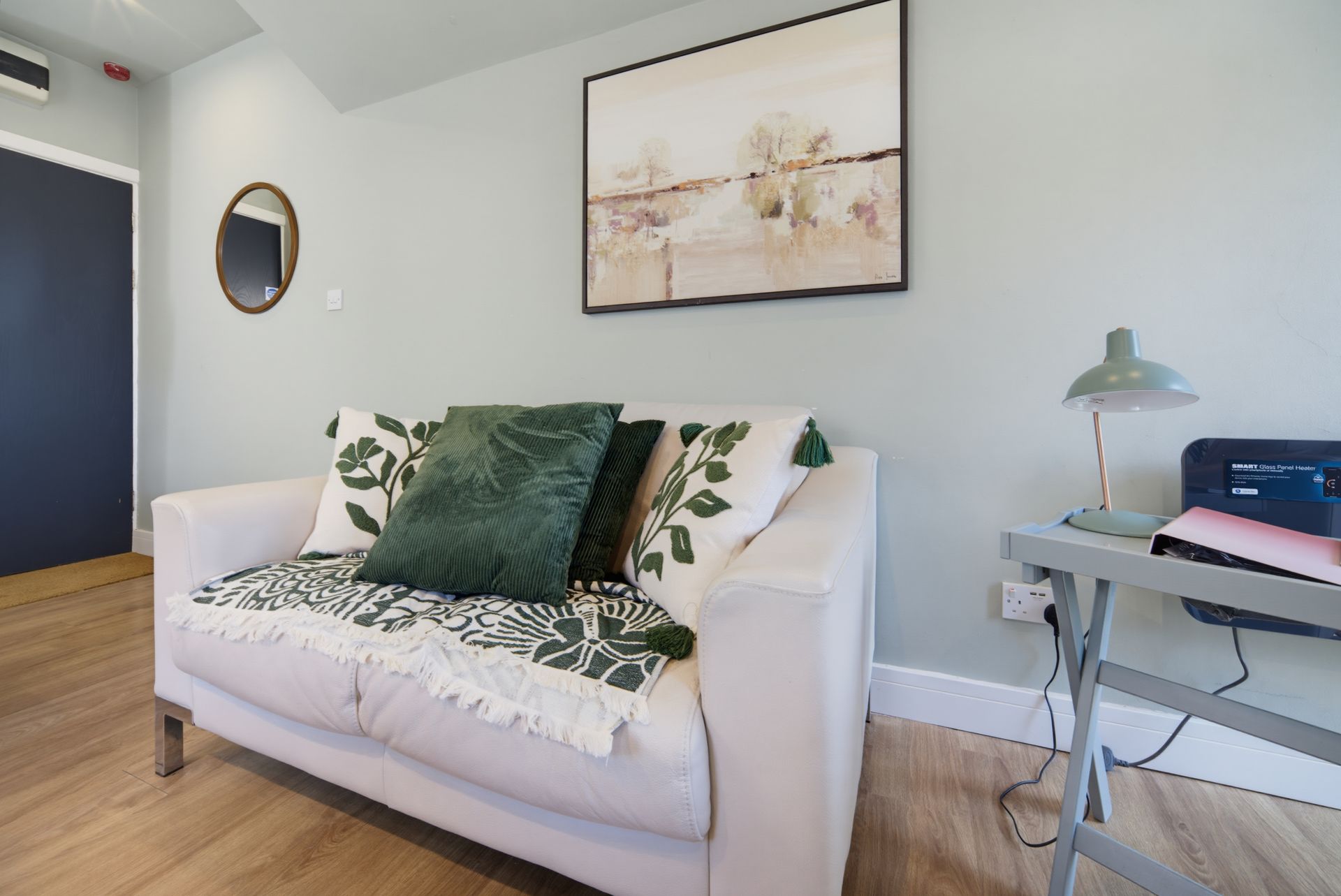 Bright living room with white sofa, green pillows, wall art, and a side table with lamp and printer.