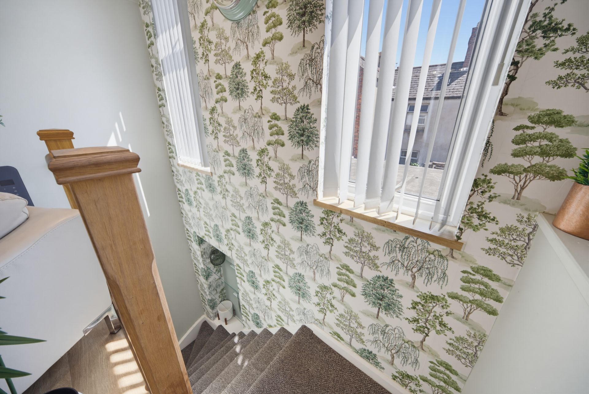Staircase landing with floral wallpaper, white curtains, and a wood banister beside a window.