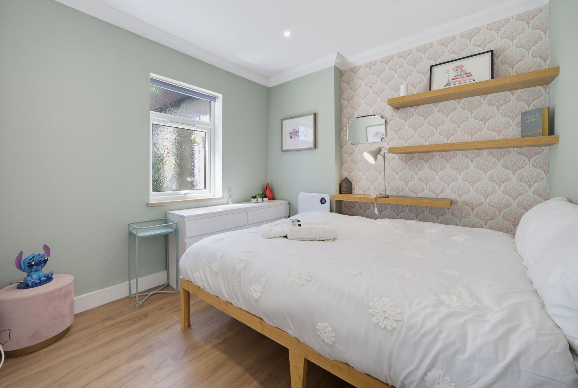 Bright bedroom with a white bed, mint walls, wooden floor, and floating shelves beside a window.