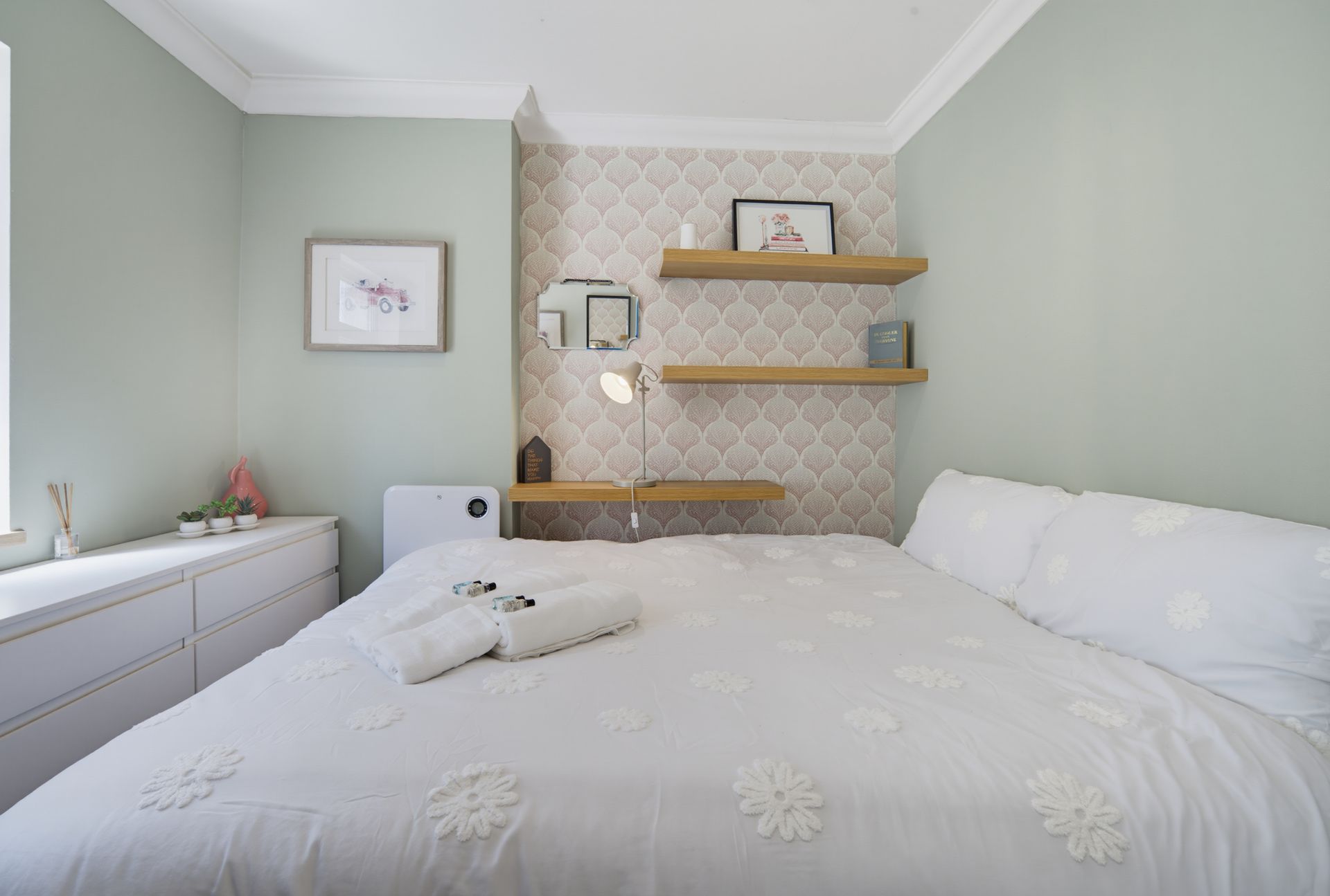 Bright bedroom with a large white bed, sage walls, dresser, and wall shelves above a patterned headboard.