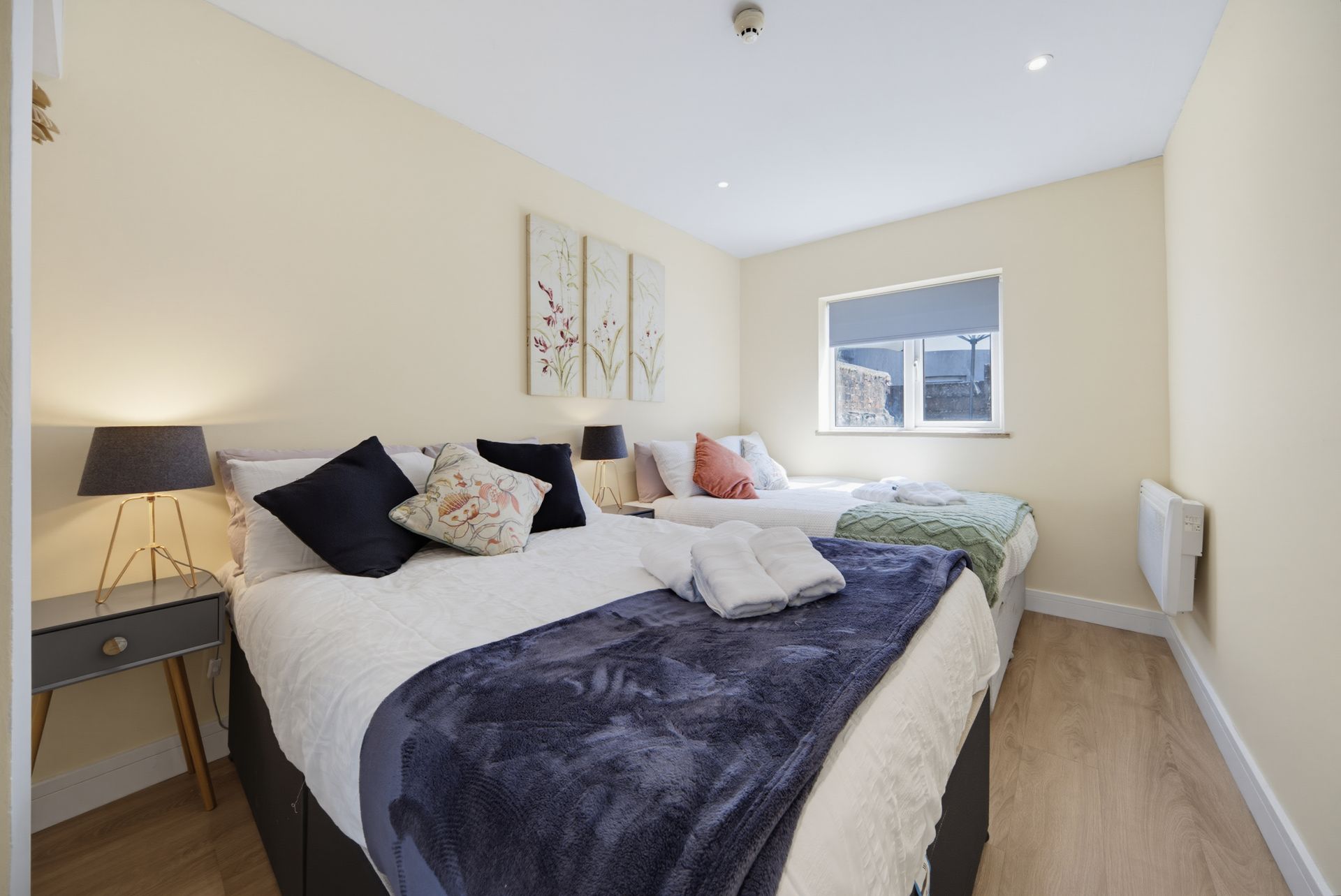 Bright bedroom with two neatly made beds, beige walls, and a window letting in daylight.