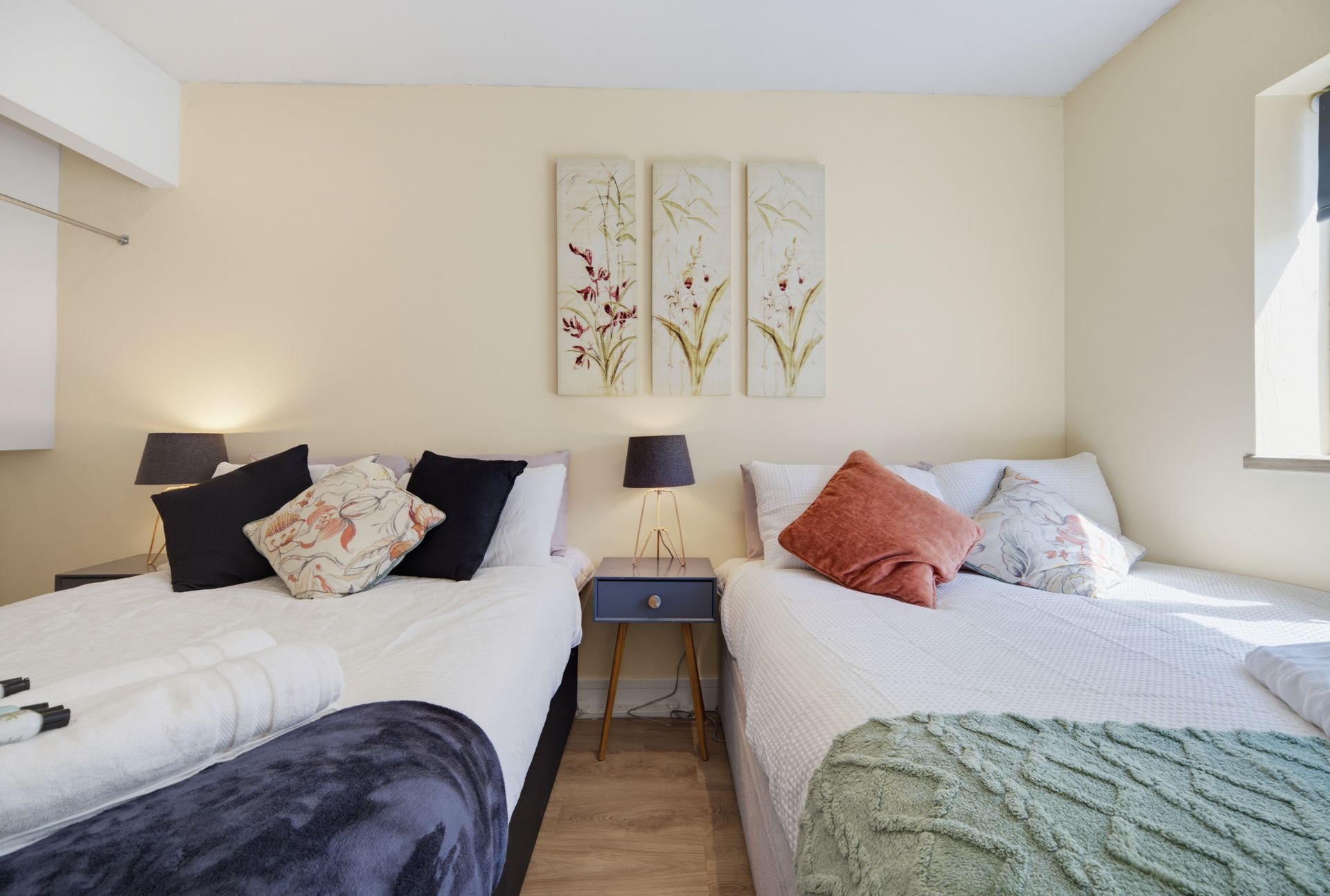 Bright bedroom with two twin beds, dark and coral pillows, and floral wall art between them.