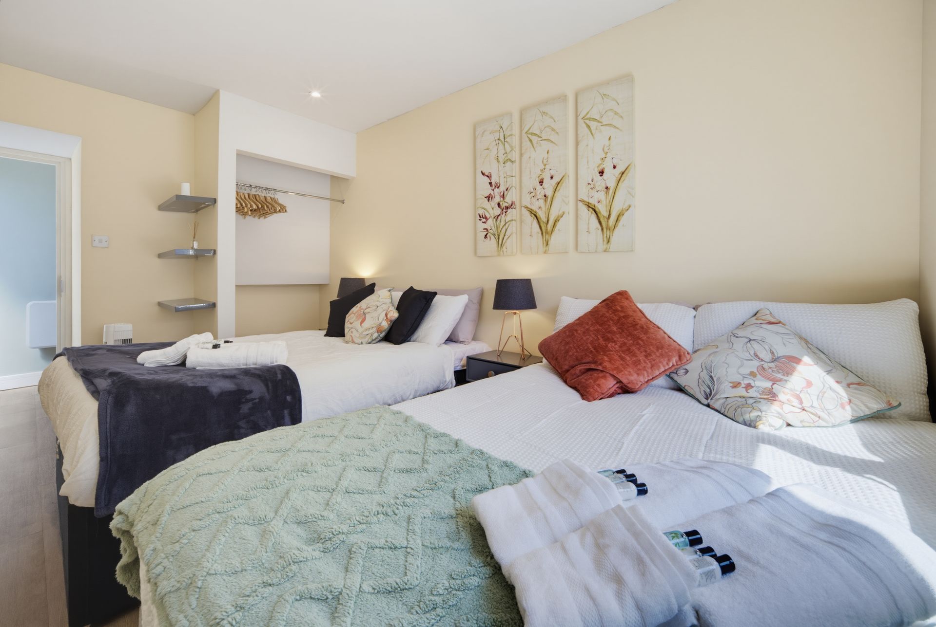 Bright bedroom with two beds, cream walls, and teal and black bedding in a sunlit hotel-style room