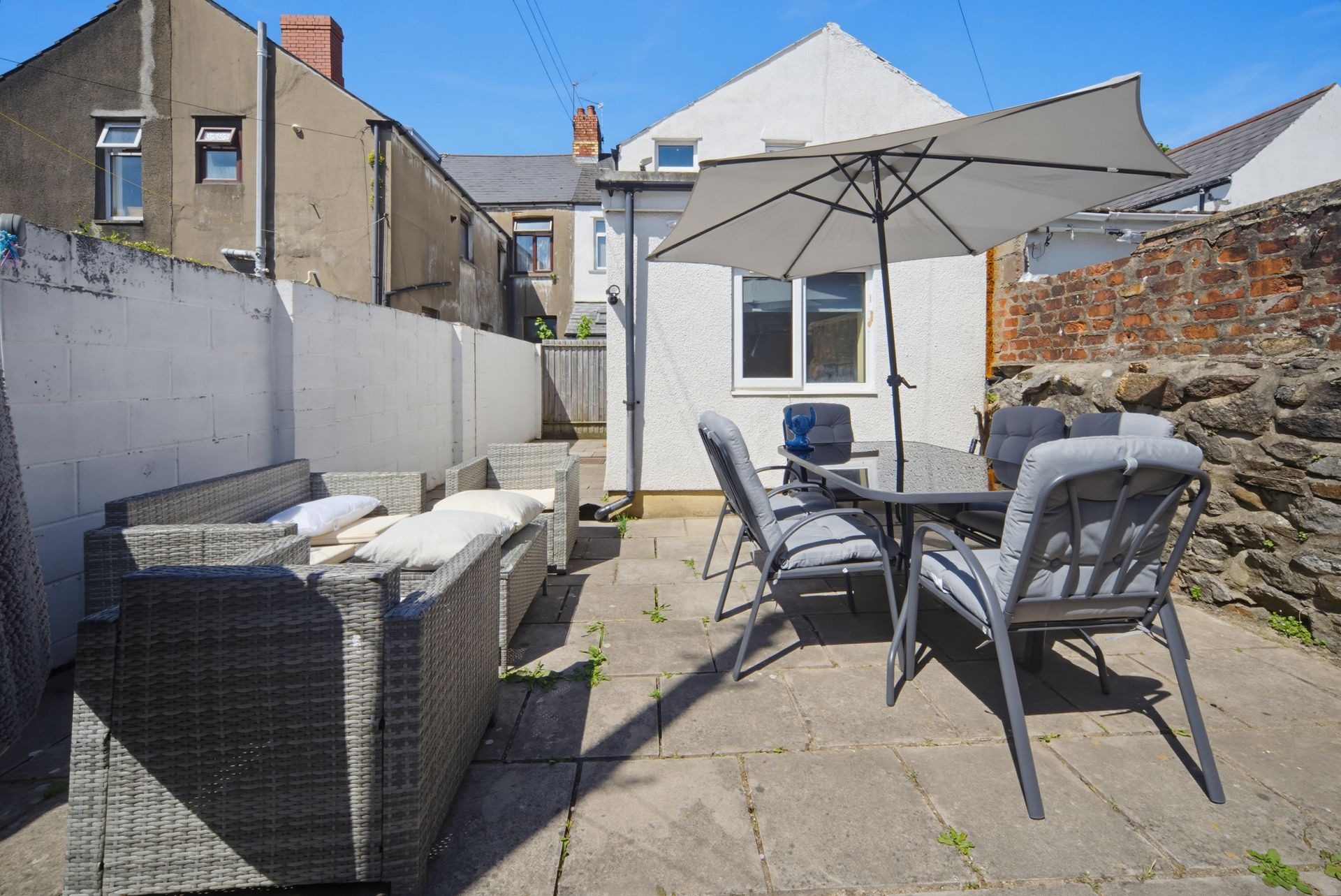 Small stone patio with outdoor chairs, table, sofa, and a white umbrella beside a house wall.
