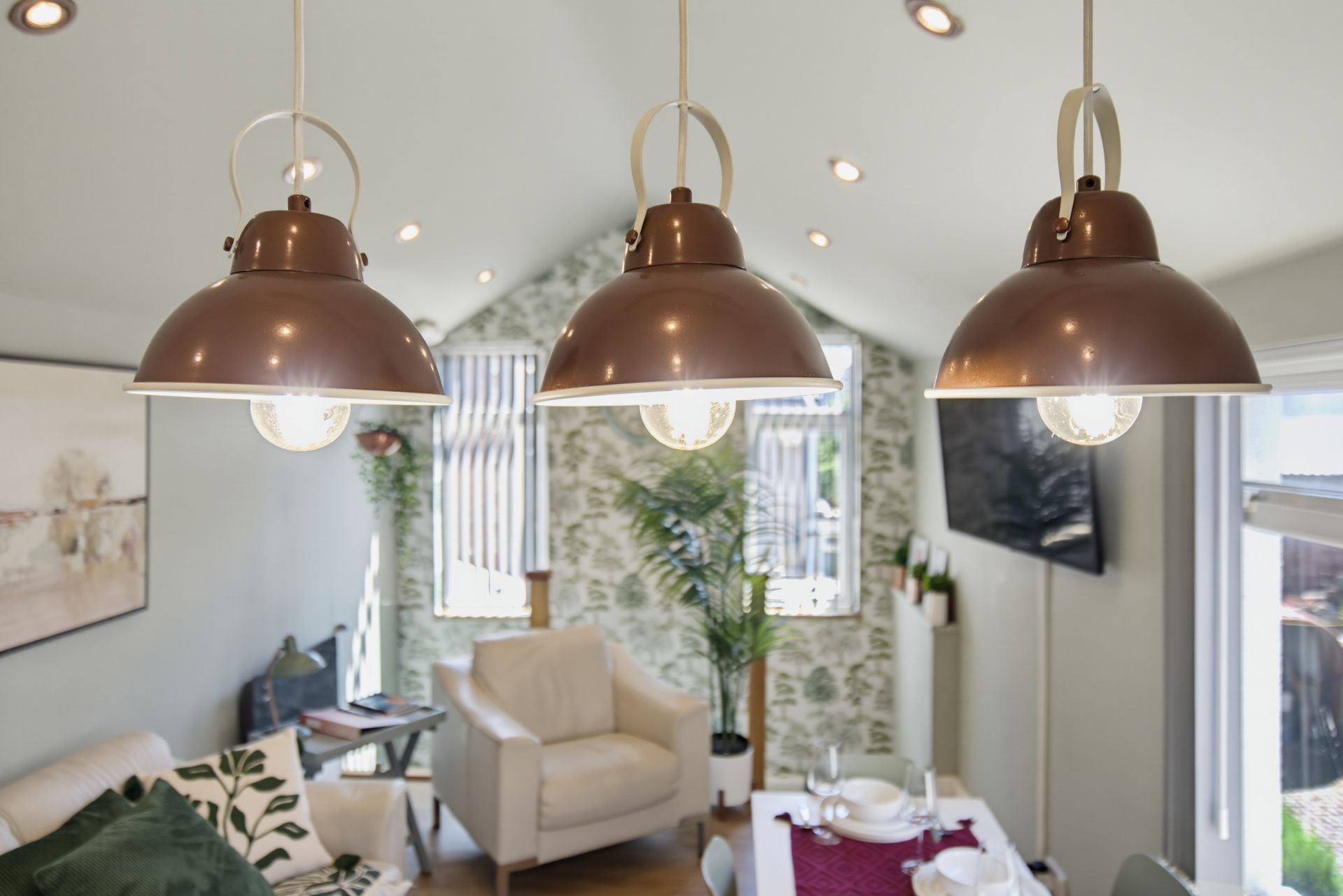 Cozy living room with three copper pendant lights, beige chair, plants, and wall-mounted TV