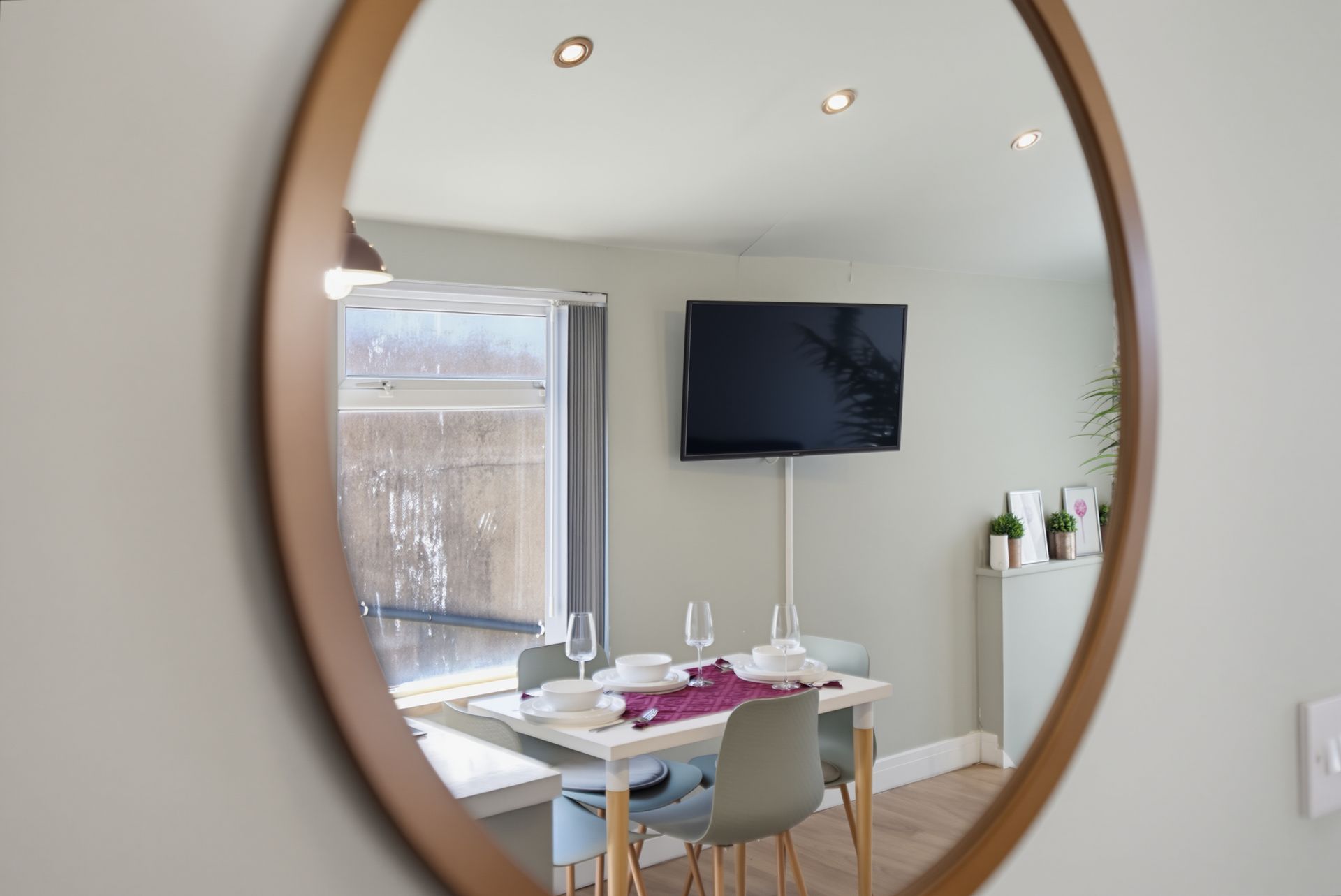 Oval mirror reflecting a bright dining area with table, chairs, wall-mounted TV, and window