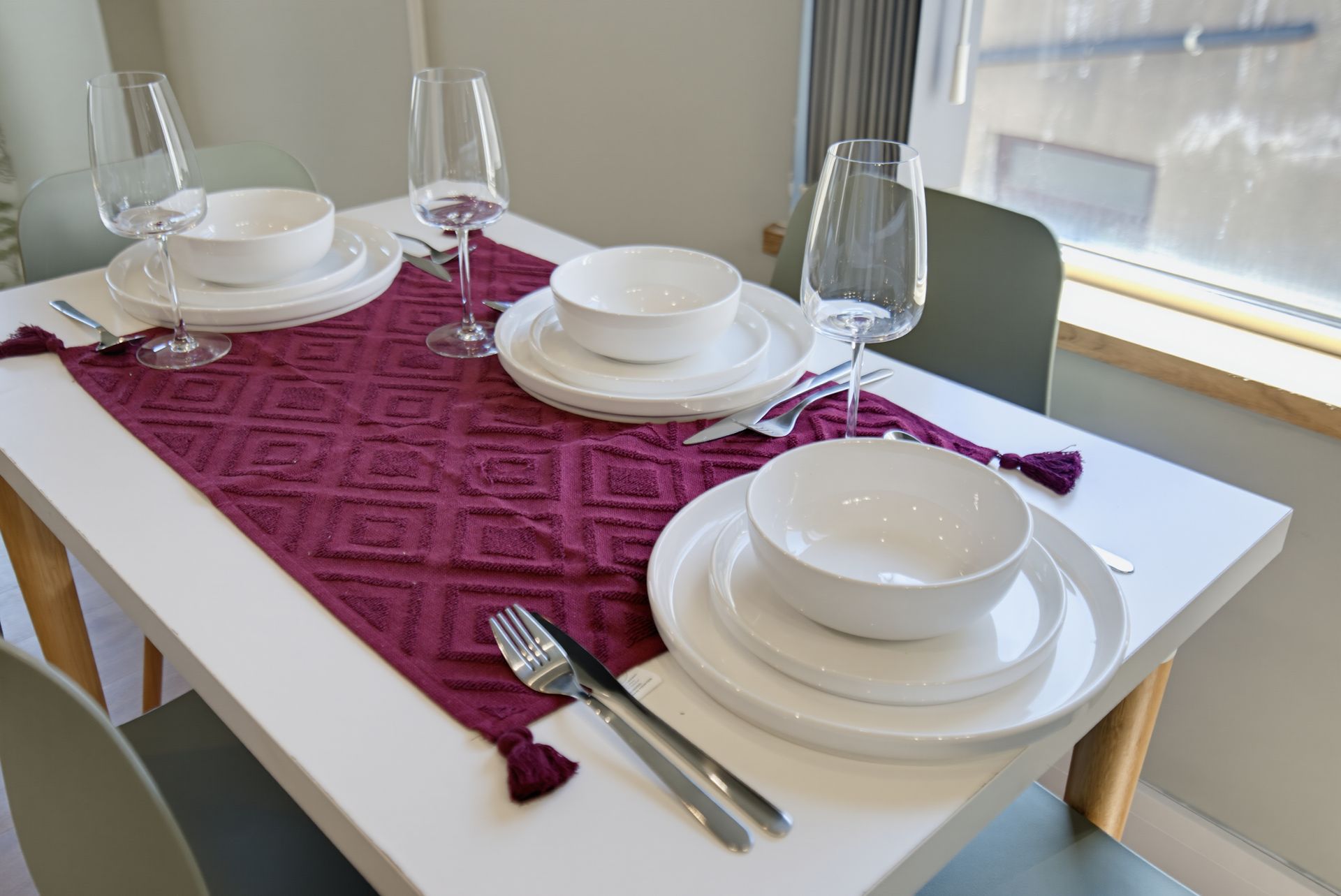 Dining table set with white dishes, glasses, and silverware on a burgundy runner near a window.