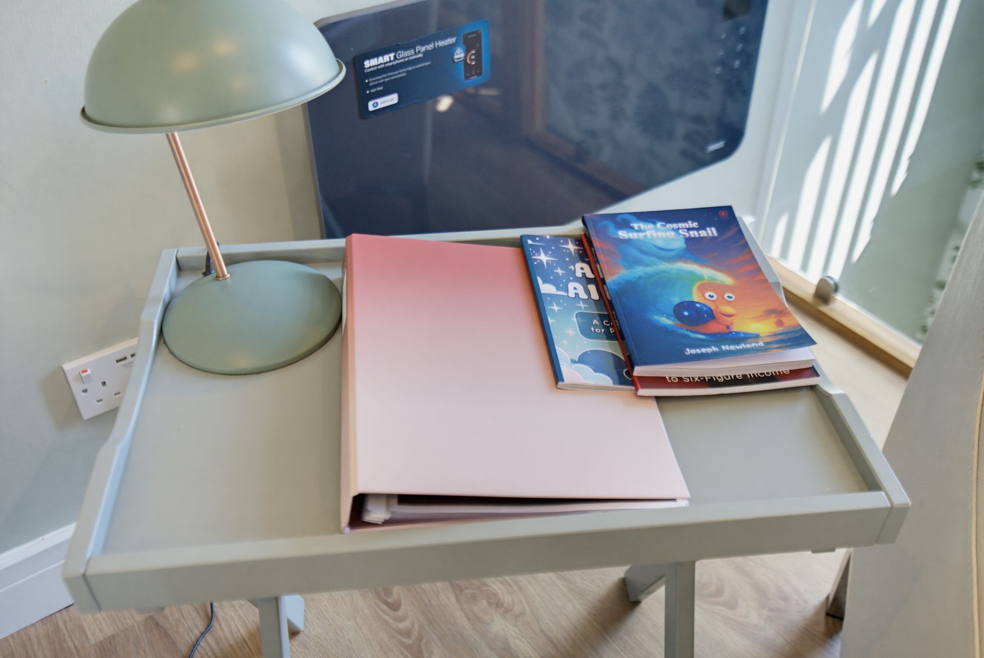 A desk with a green lamp, a pink notebook, and two books beside a window.