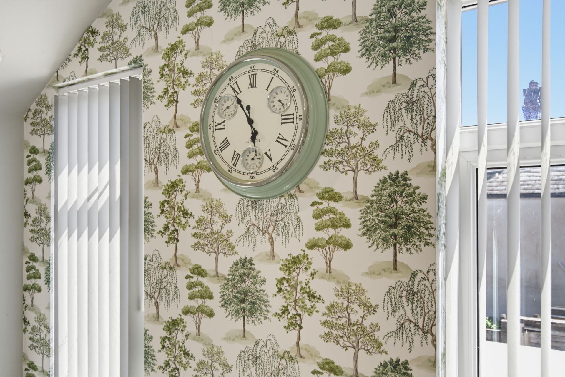 Green tree-pattern wallpaper with a vintage wall clock between two windows with white blinds