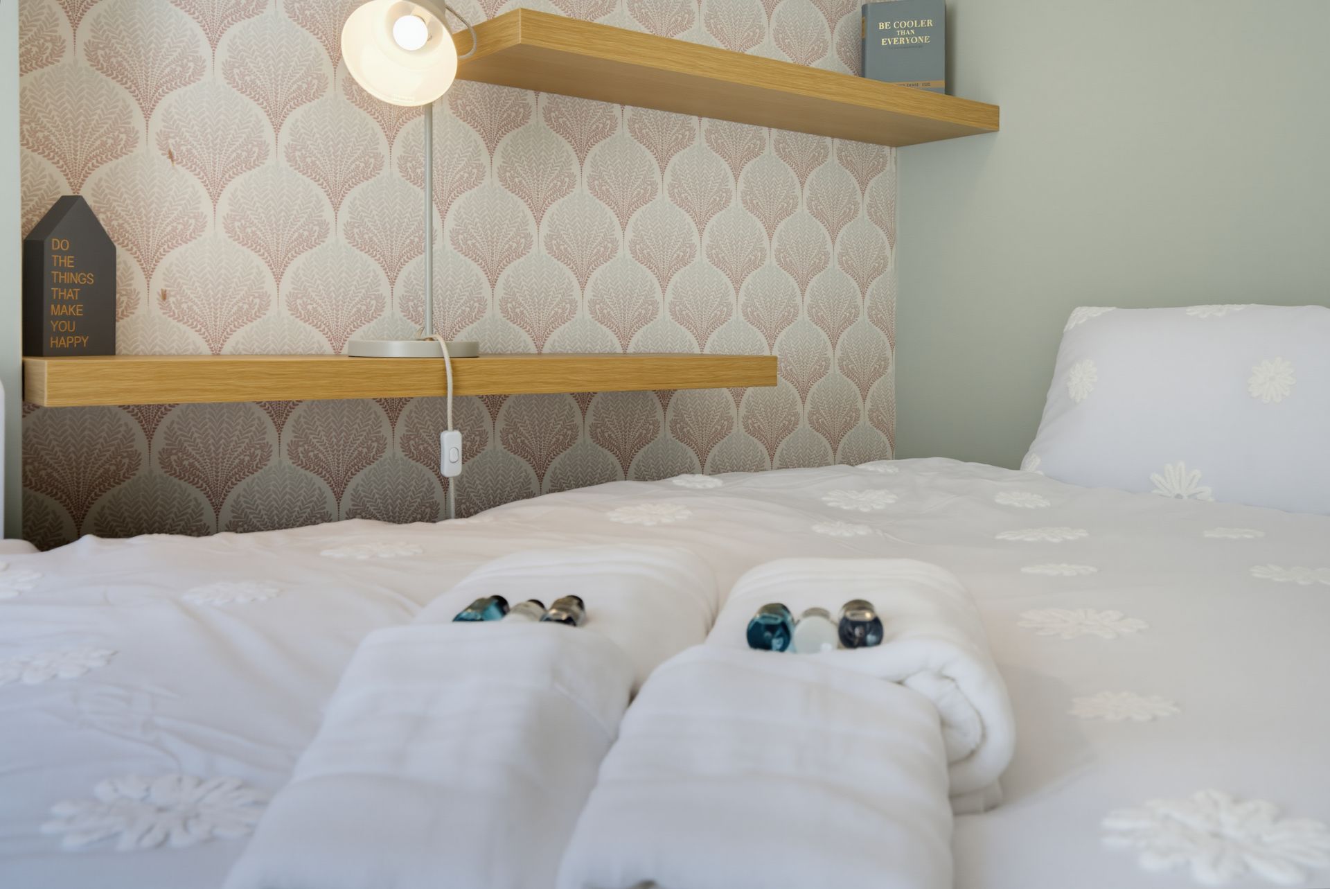 Neatly made white hotel bed with rolled towels and a bedside shelf lamp against patterned wallpaper