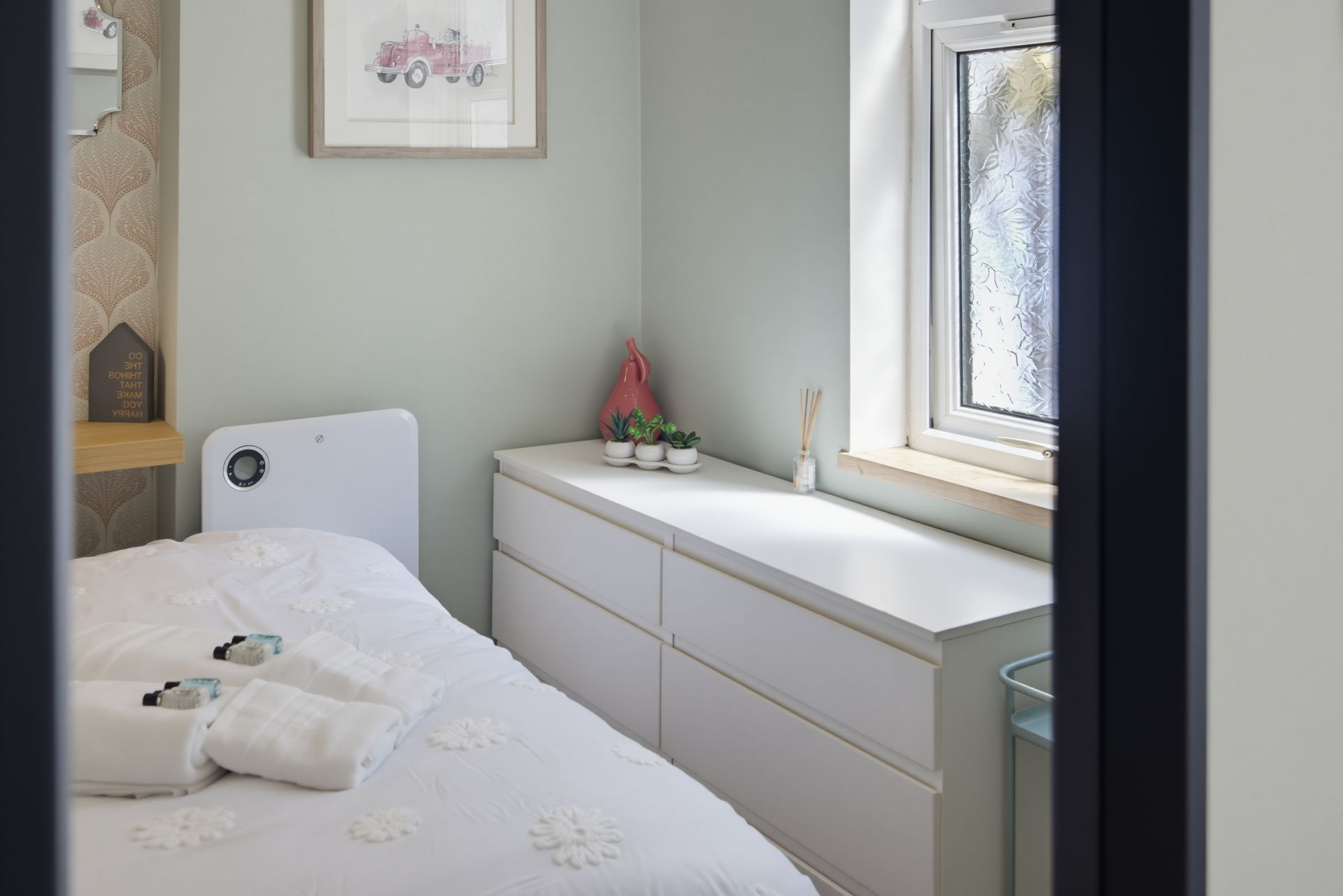 Small bedroom with white bed, dresser, window, and soft neutral decor.