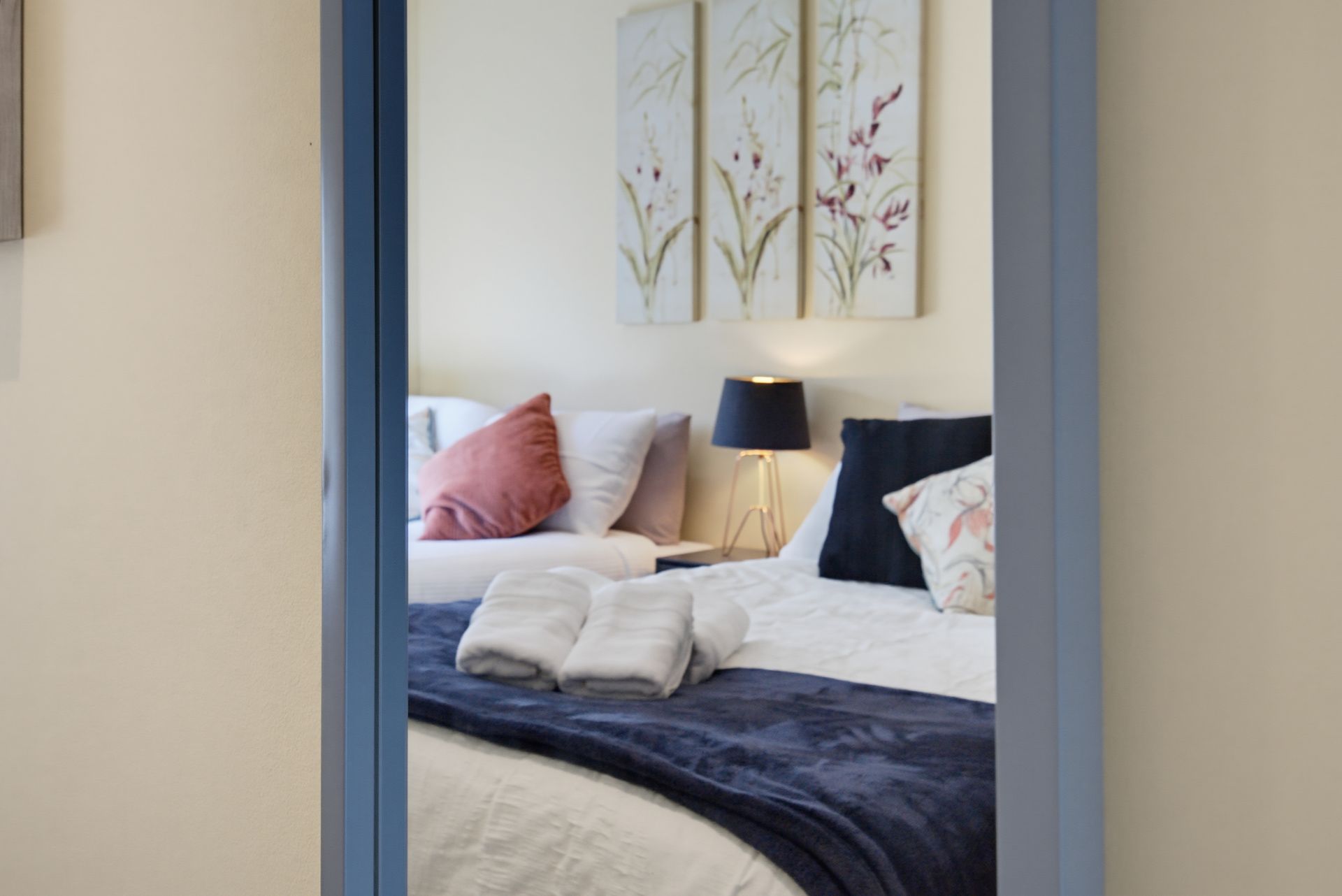 Blue-trimmed mirror reflecting a cozy bedroom with a bed, pillows, bedside lamp, and floral wall art