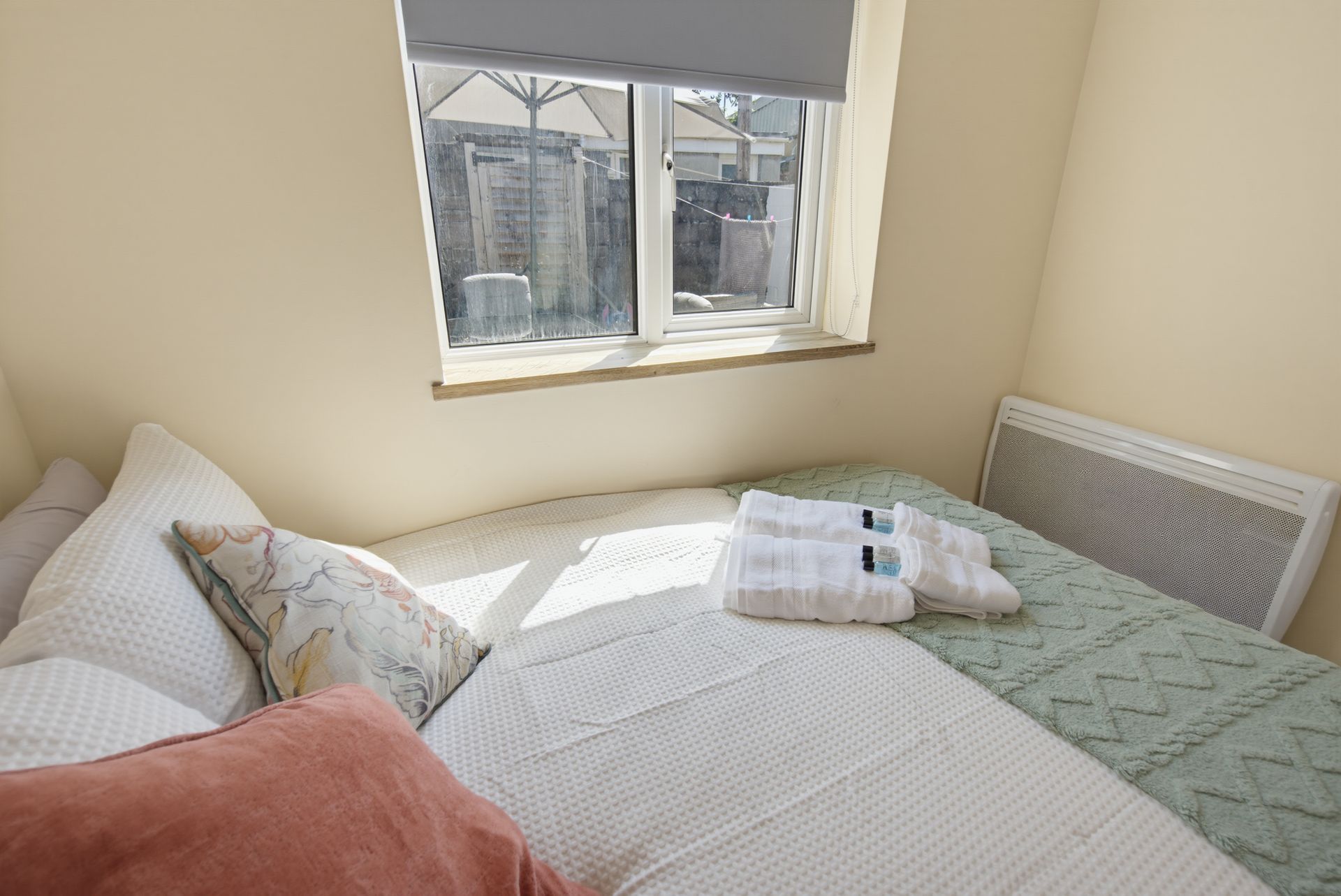 Neatly made bed by a bright window in a beige bedroom, with folded towels and pillows.