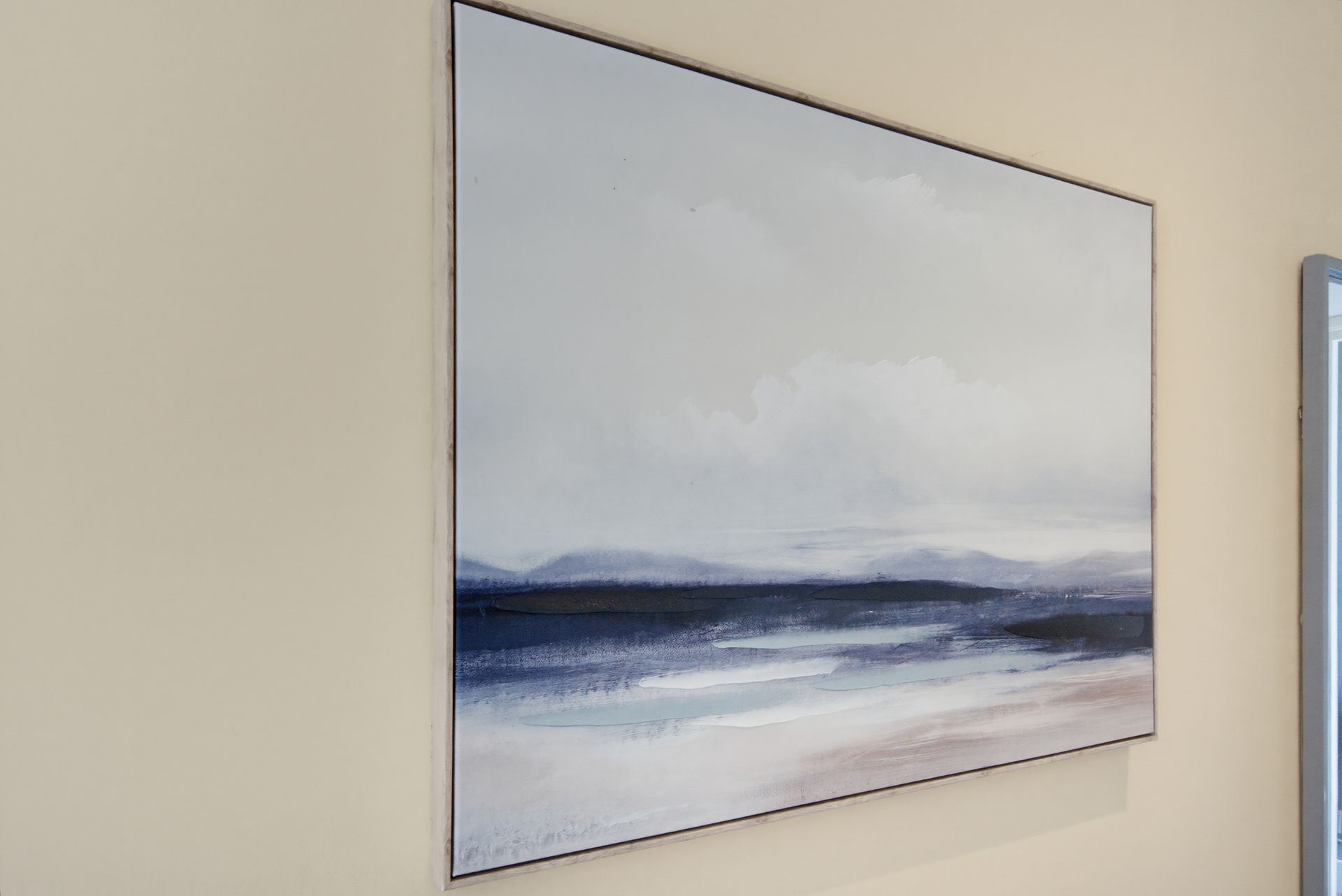 Framed minimalist seascape painting in soft blue and gray tones hanging on a beige wall