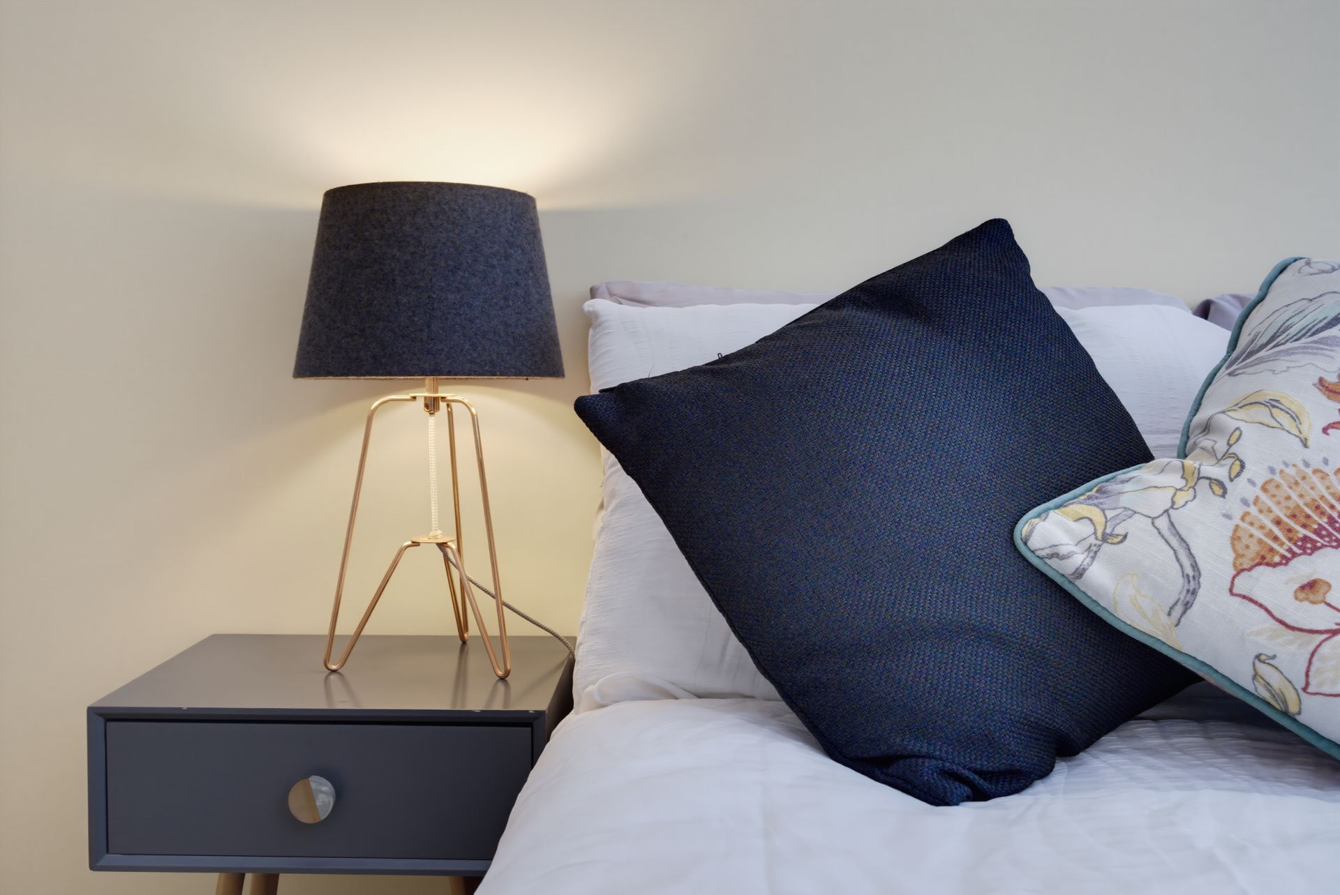 Small bedside table with a dark lamp beside a white bed with navy and floral pillows