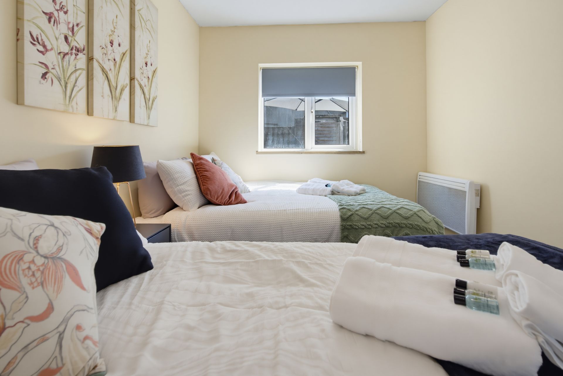 Neatly made bedroom with two beds, floral wall art, and a window above the far bed.