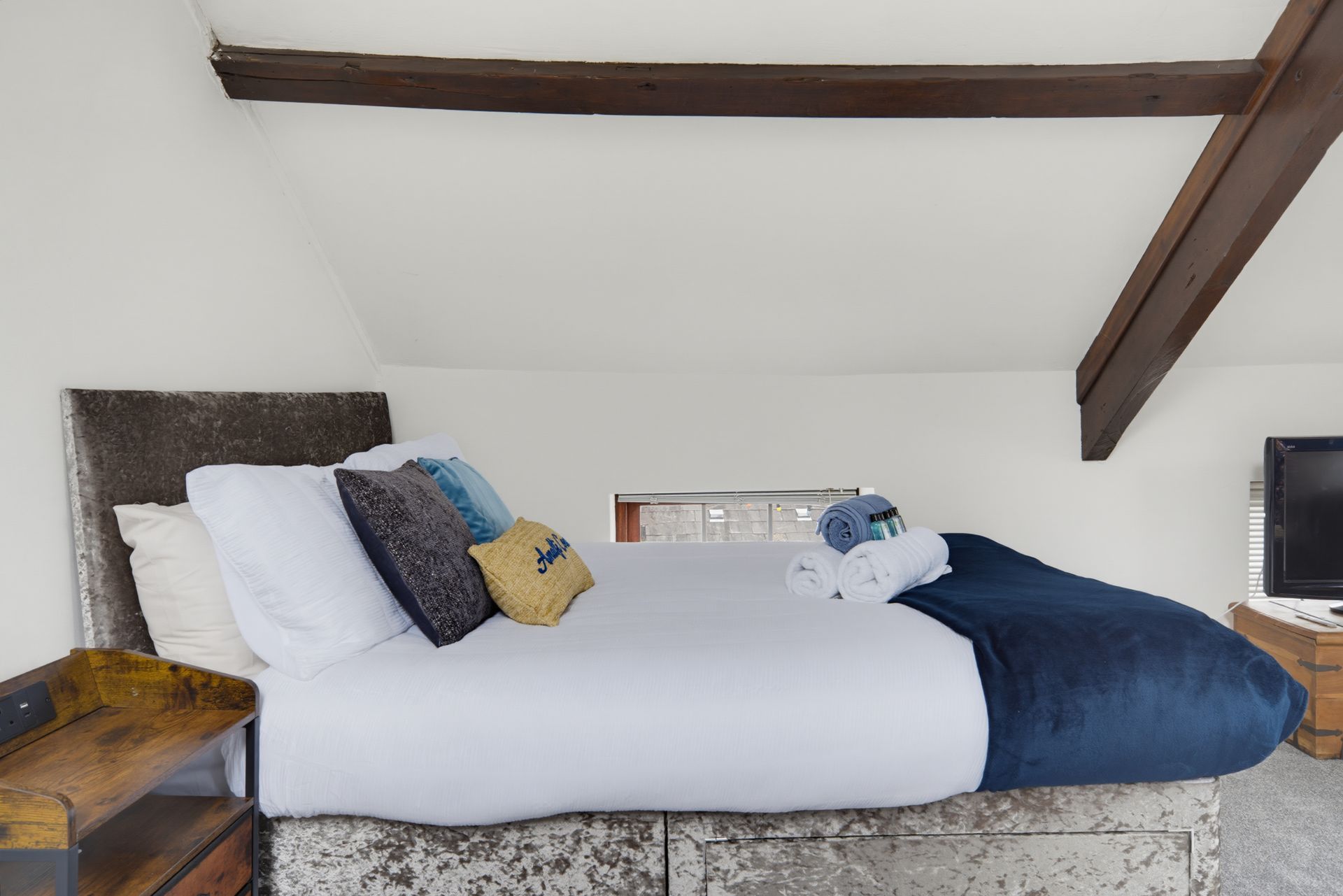 Bedroom with a made bed, white bedding, blue throw, pillows, and exposed wooden beams above.