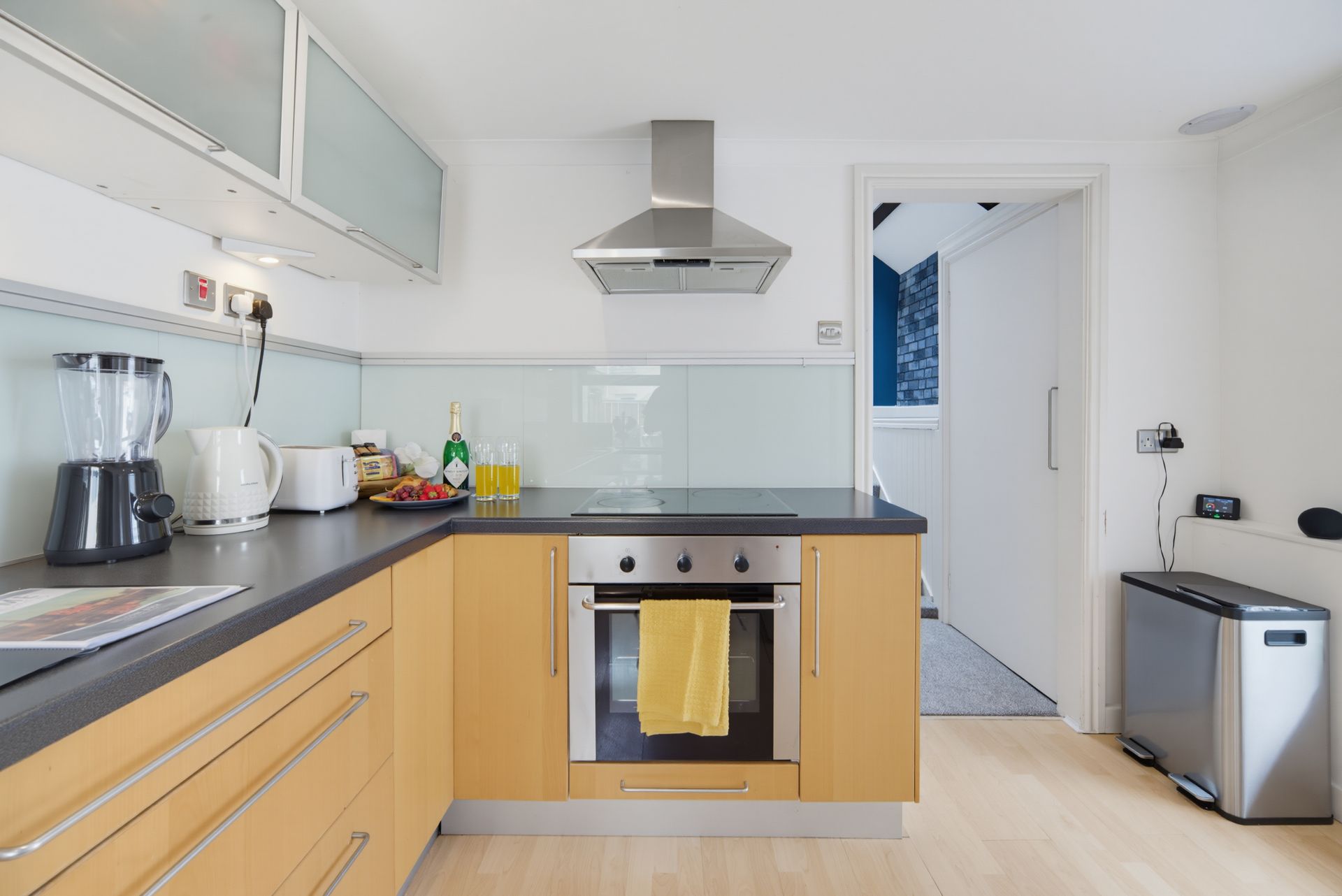 Modern kitchen with light wood cabinets, black countertops, stainless range hood, and a small trash bin.