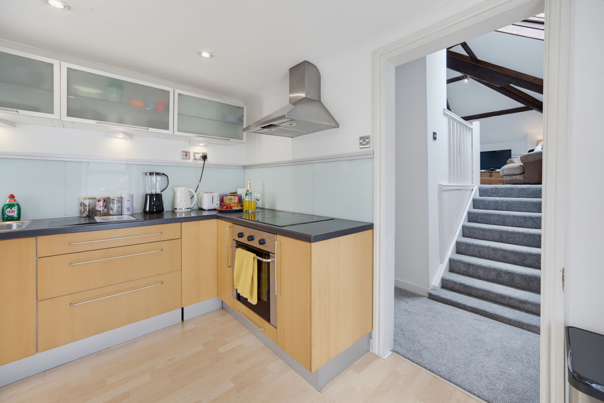 Modern kitchen with light wood cabinets, gray countertops, and a doorway to a carpeted staircase.
