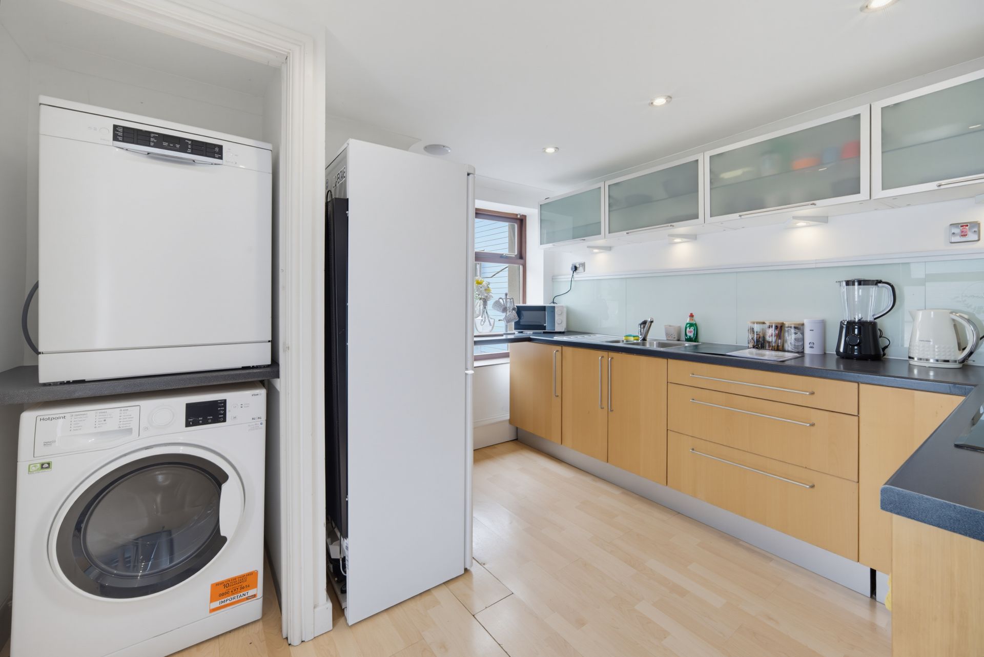 Bright modern kitchen with light wood cabinets, white appliances, and stacked washer and dryer.