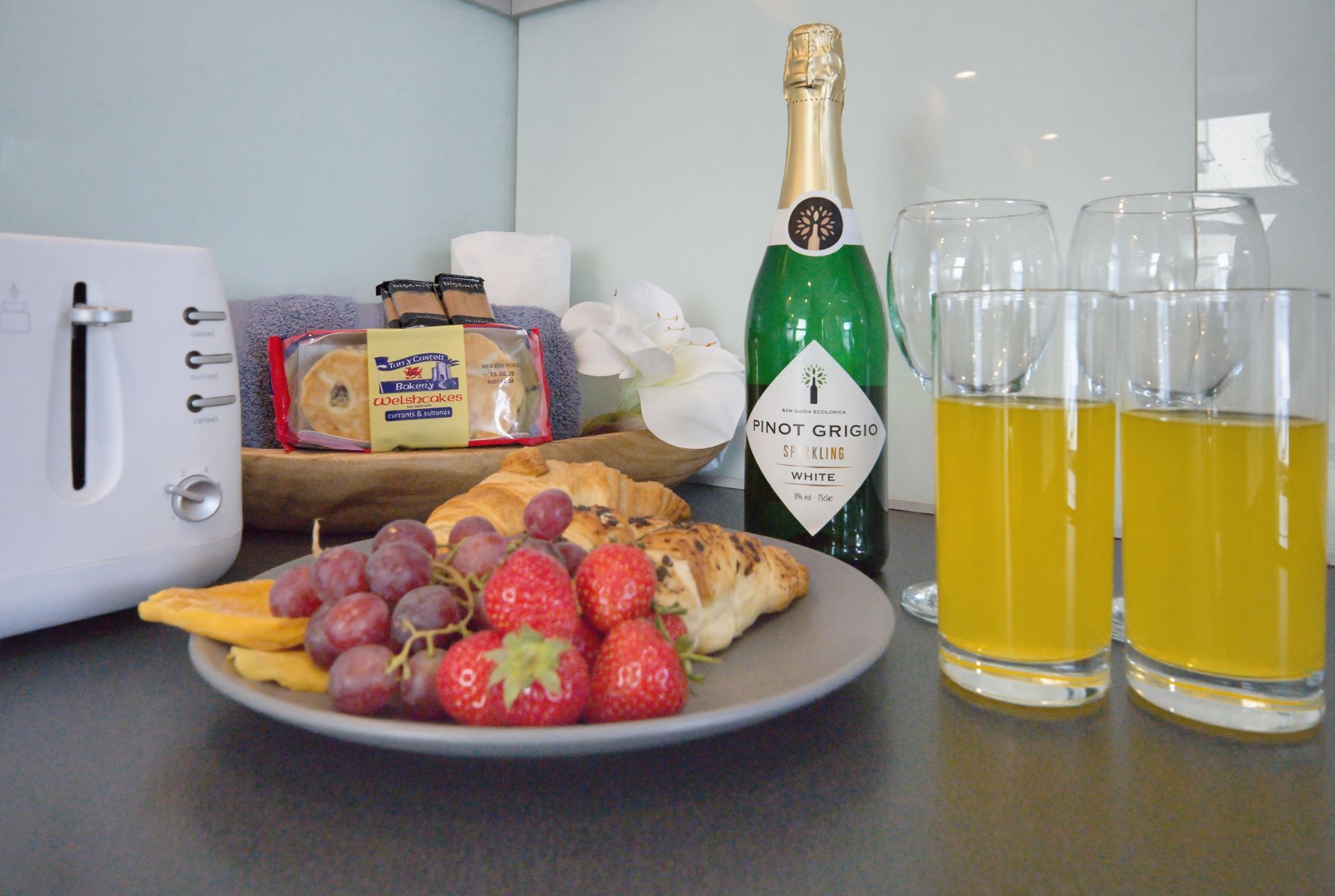 Breakfast spread with fruit, pastries, champagne, and two glasses of orange juice on a kitchen counter