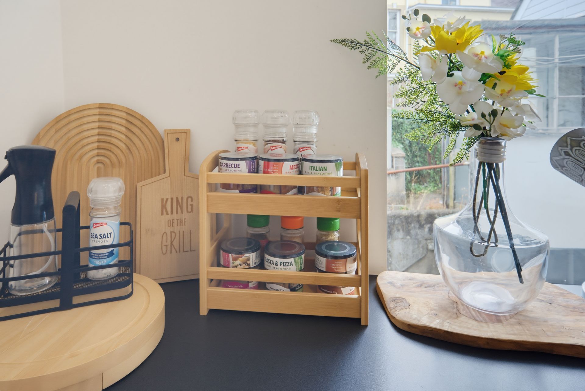 Kitchen countertop with spice rack, jars, cleaning spray, and white-yellow flowers in a vase by a window