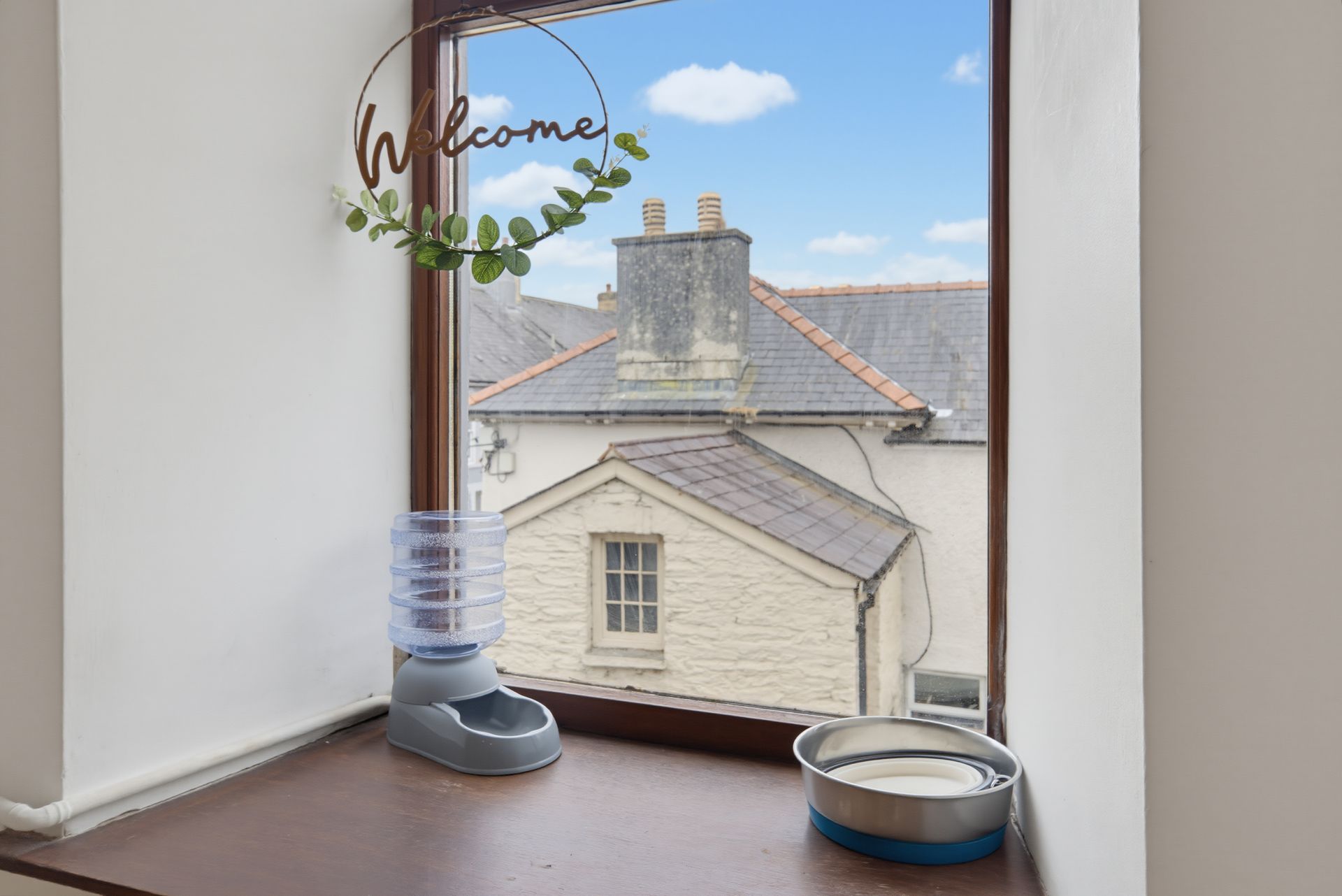 Window nook with a “Welcome” hanging sign, blue diffuser, and pet water bowl overlooking rooftops