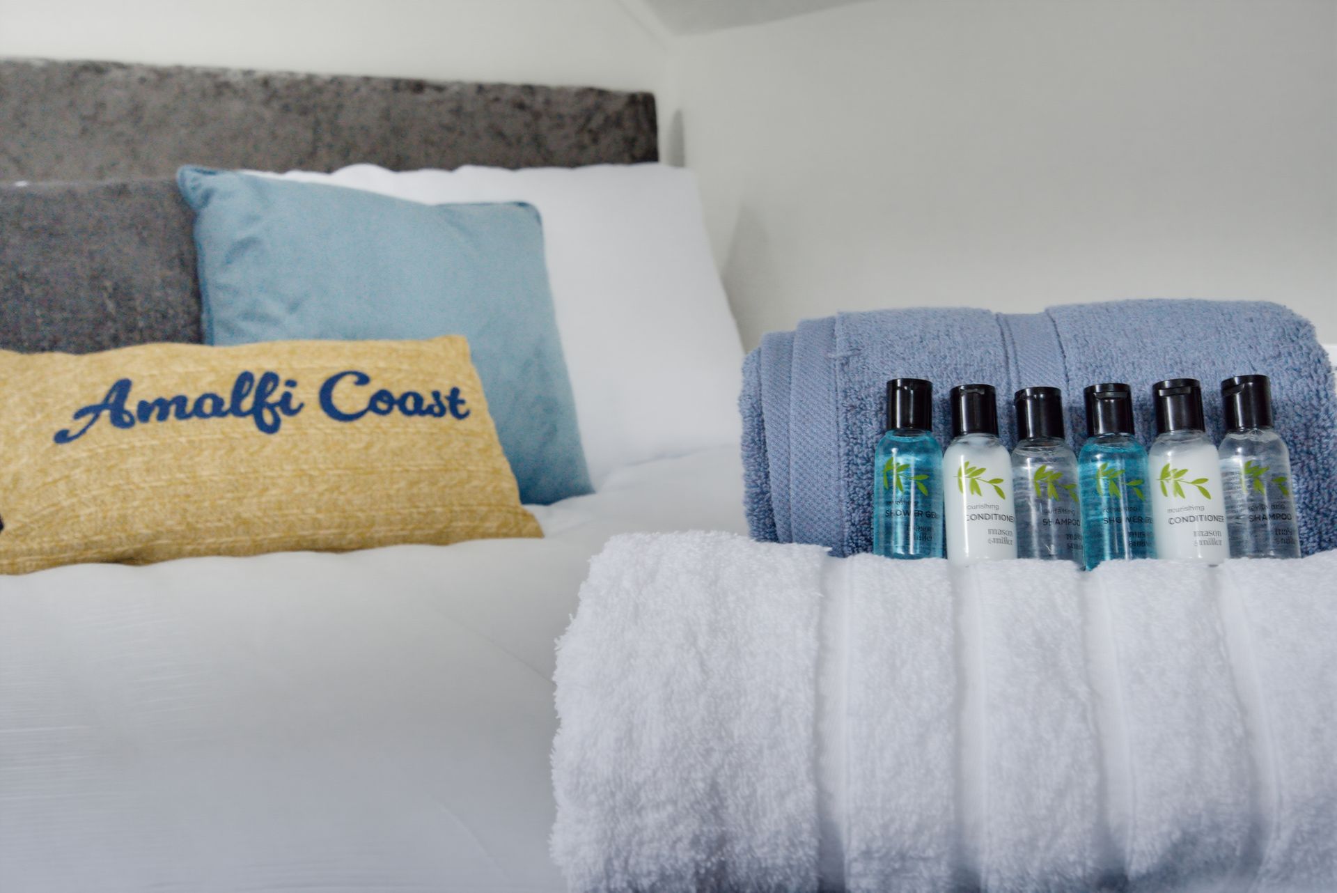 Bed with pillows and a tray holding small bottles on a white blanket, with an “Amalfi Coast” pillow in front