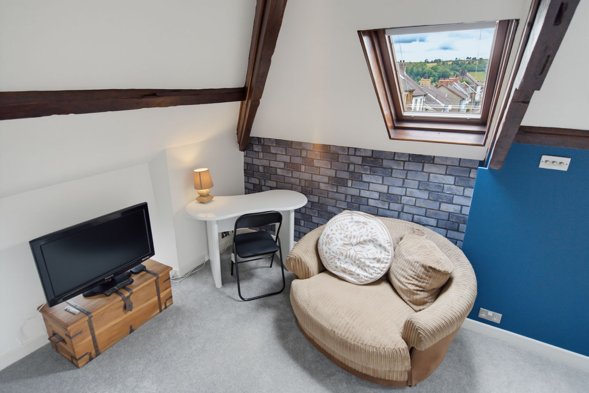 Small attic room with TV, desk, window seat, round chair, and blue accent wall