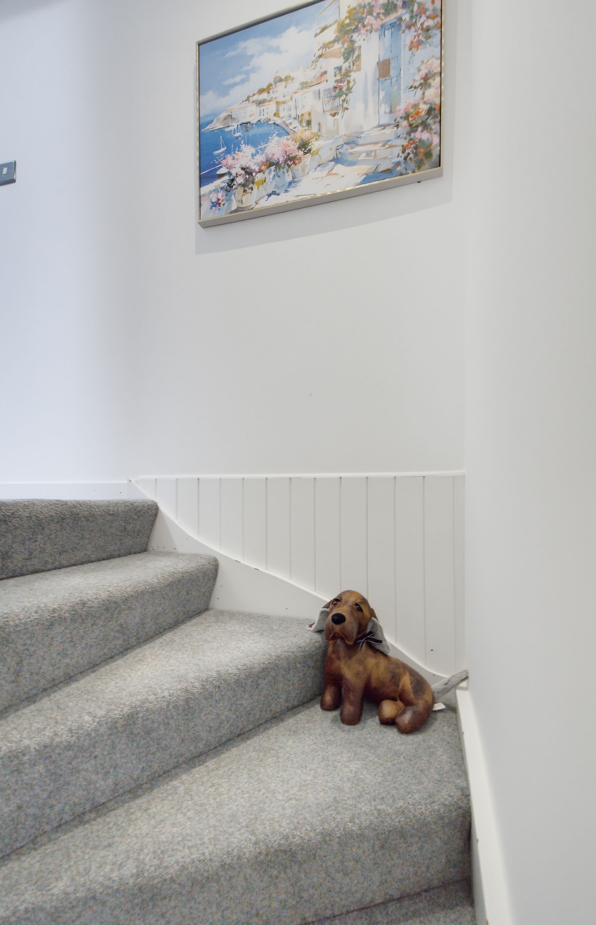Small brown dog sitting on gray carpeted stairs beside white wainscoting and a framed wall painting