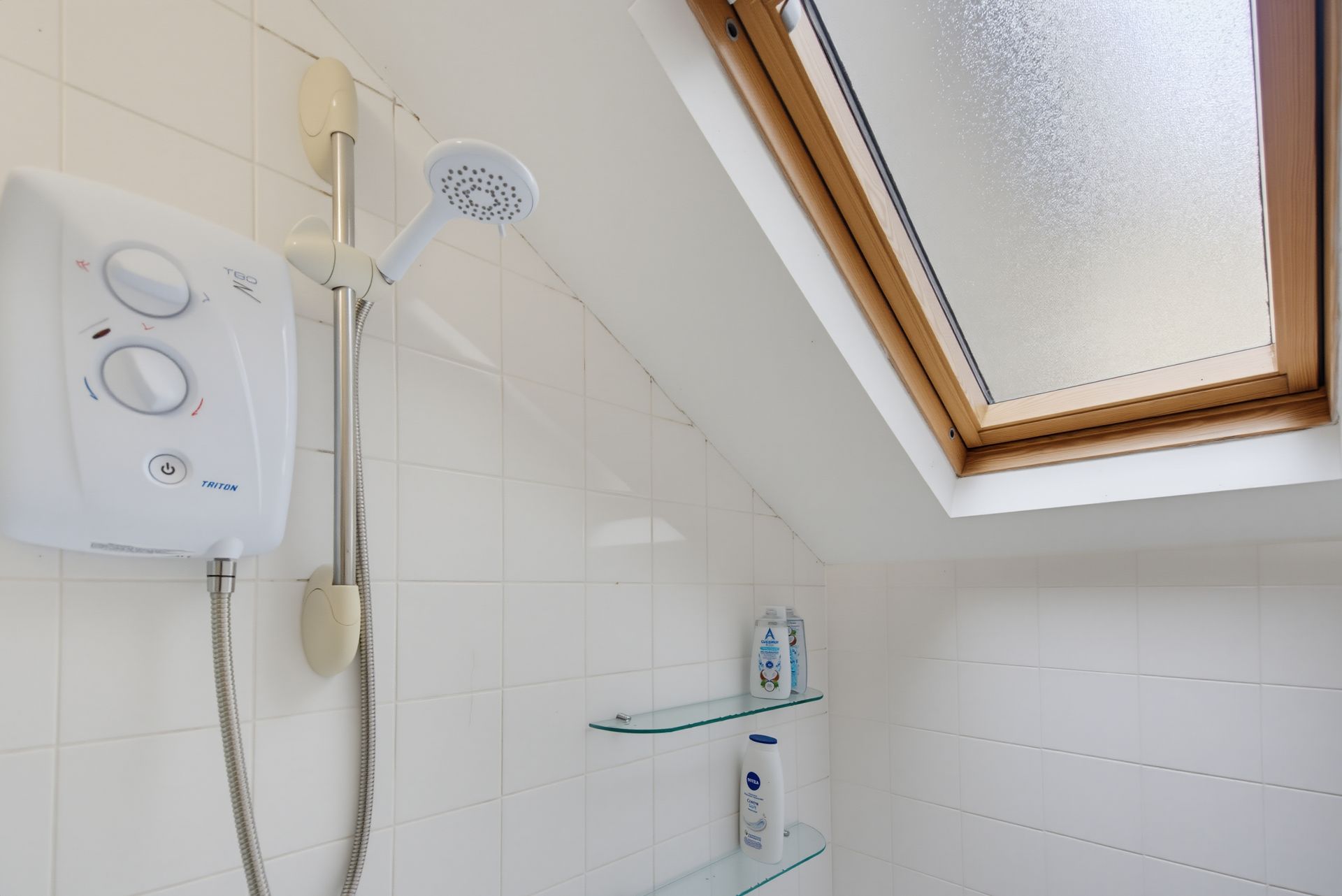 Bathroom shower with white tiled walls, wall-mounted electric shower, glass shelf, and skylight window.