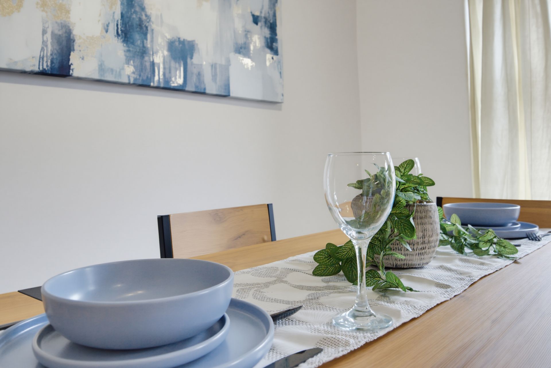 Set dining table with plates, wine glass, greenery centerpiece, and blue bowls in a bright room