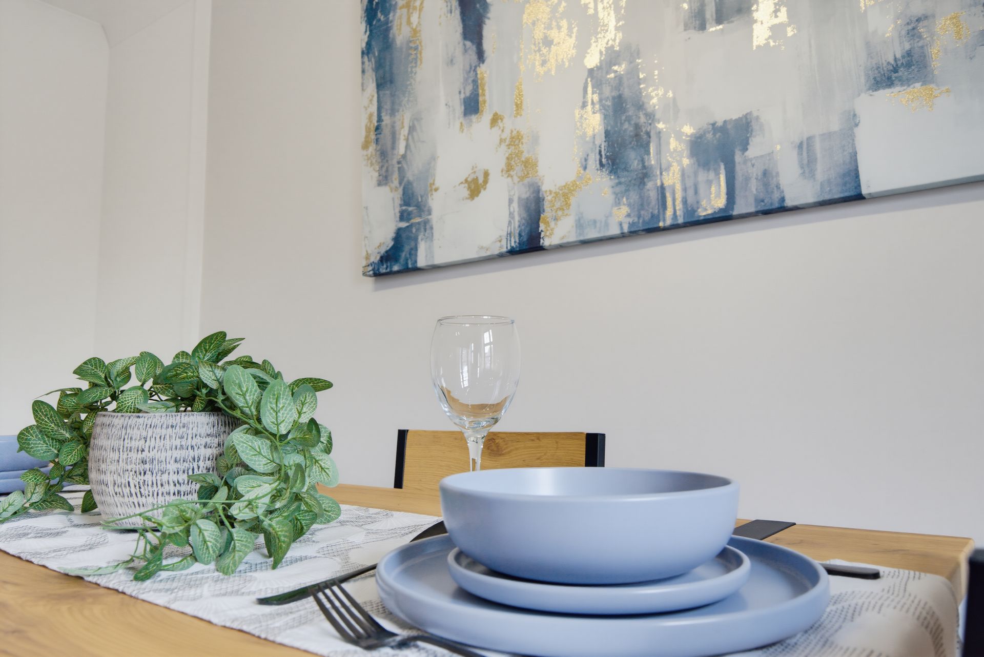 Blue dishware on a dining table with a leafy centerpiece and abstract wall art behind it