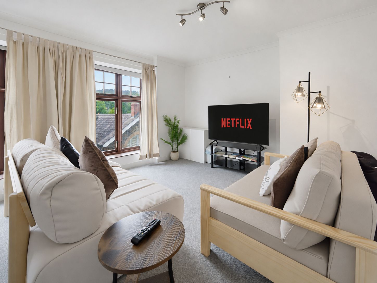 Bright living room with white sofas, a TV showing Netflix, and large windows with curtains.