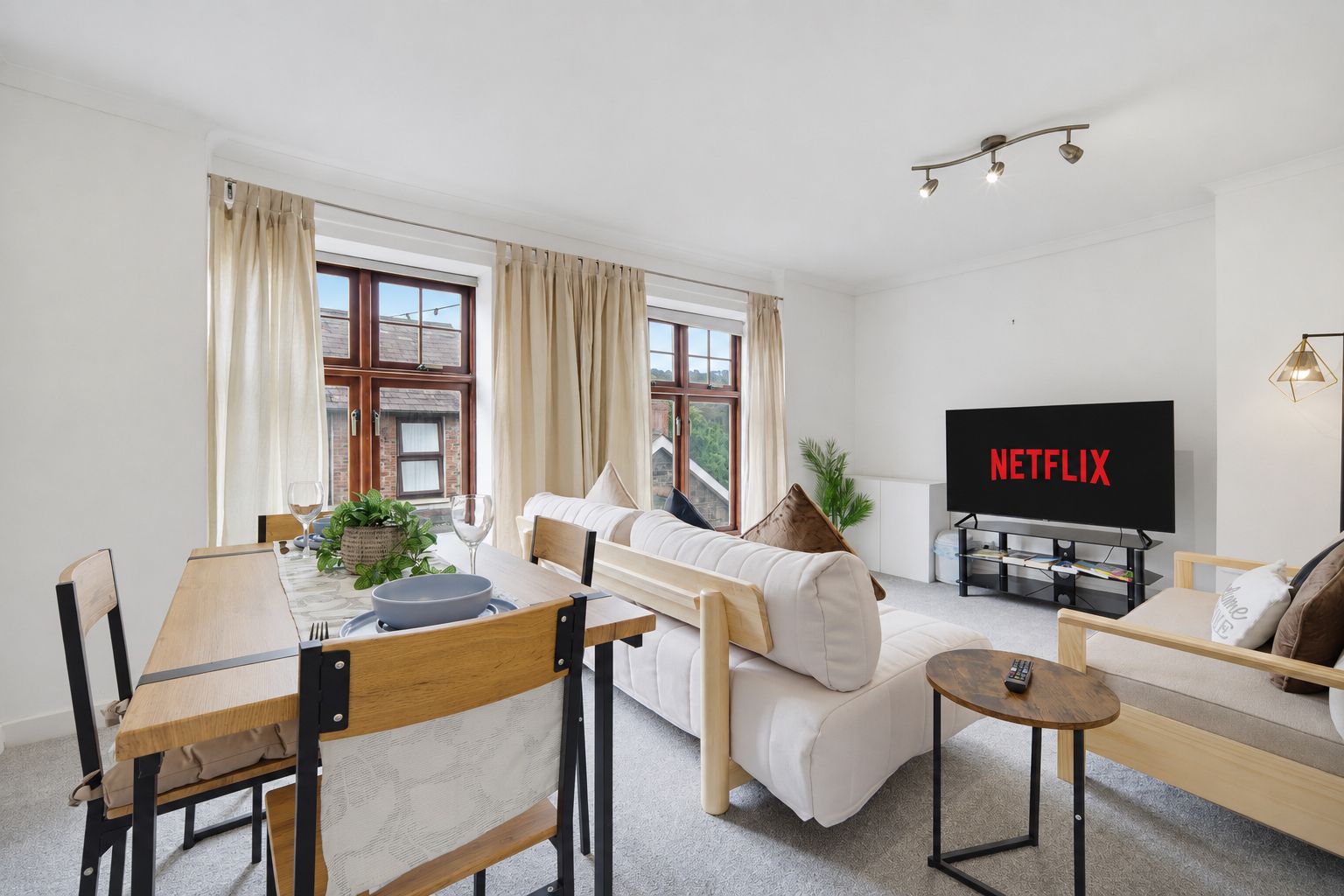 Bright living room with beige sofa, dining table, large windows, and TV displaying Netflix