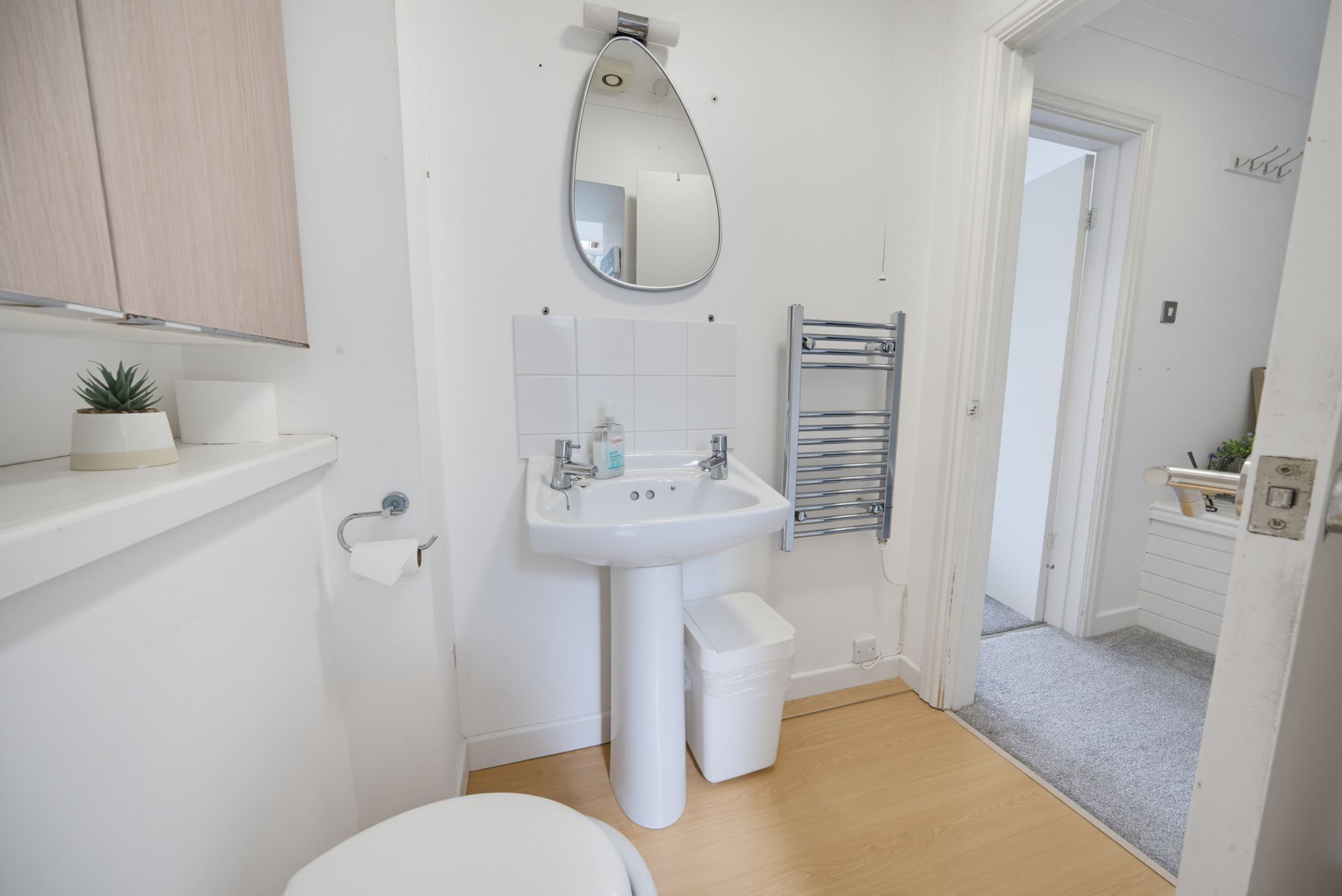 Bright white bathroom with pedestal sink, oval mirror, towel rack, and doorway to a carpeted room