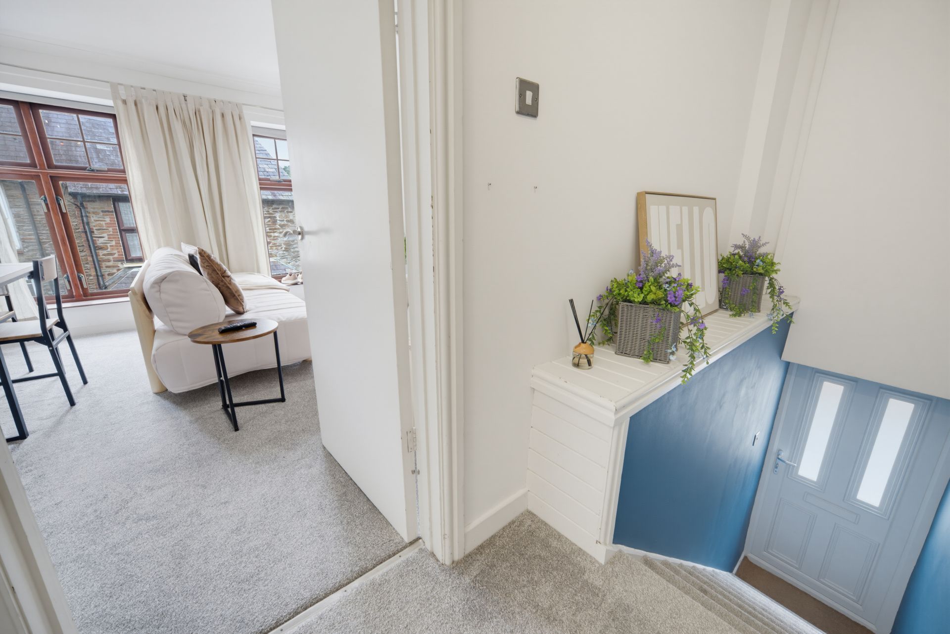 Bright hallway with white walls, gray carpet, and stairs leading down beside a small shelf with plants.