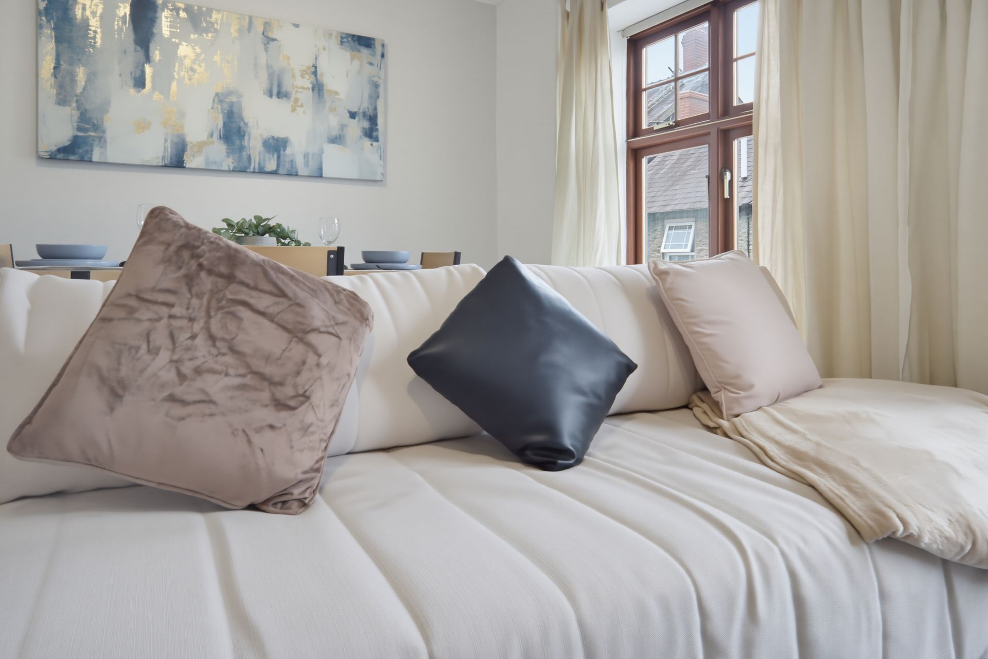 Neatly made bed with cream bedding and three decorative pillows beside a window and abstract wall art.