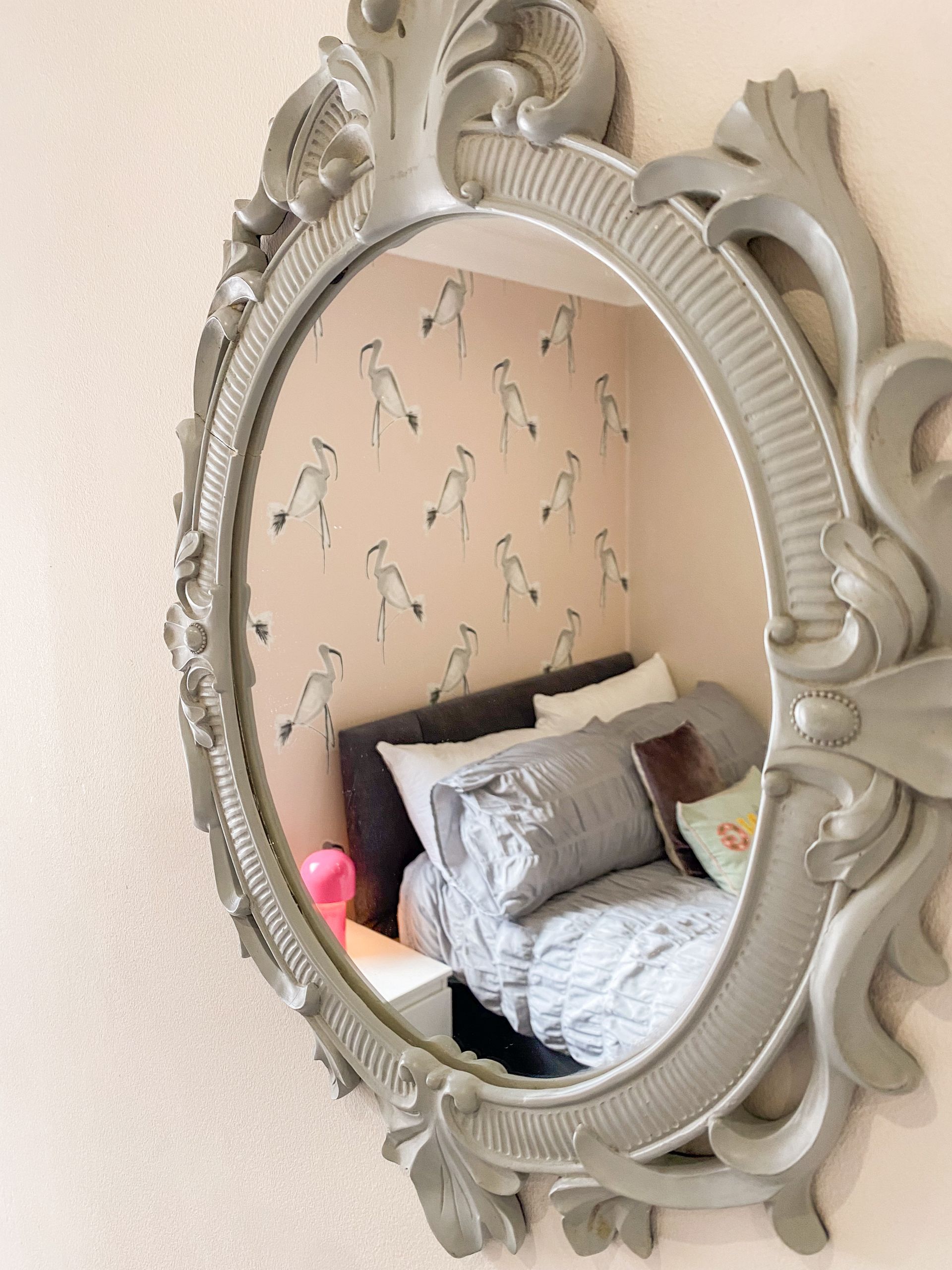 Ornate oval mirror reflecting a bed with gray pillows and pink lamp in a softly patterned bedroom