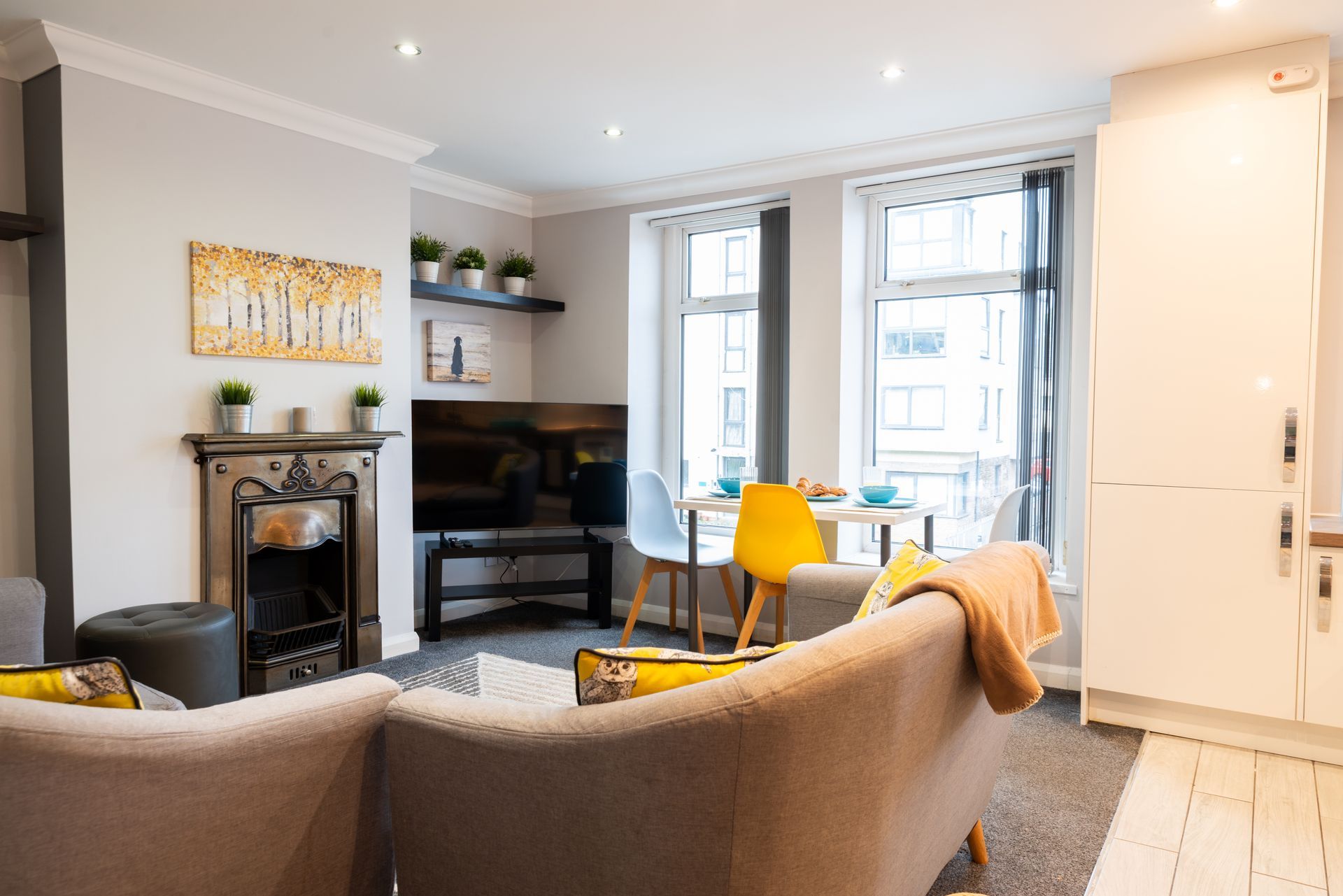 Bright modern living room with sofa, piano, fireplace, and yellow chairs by tall windows.
