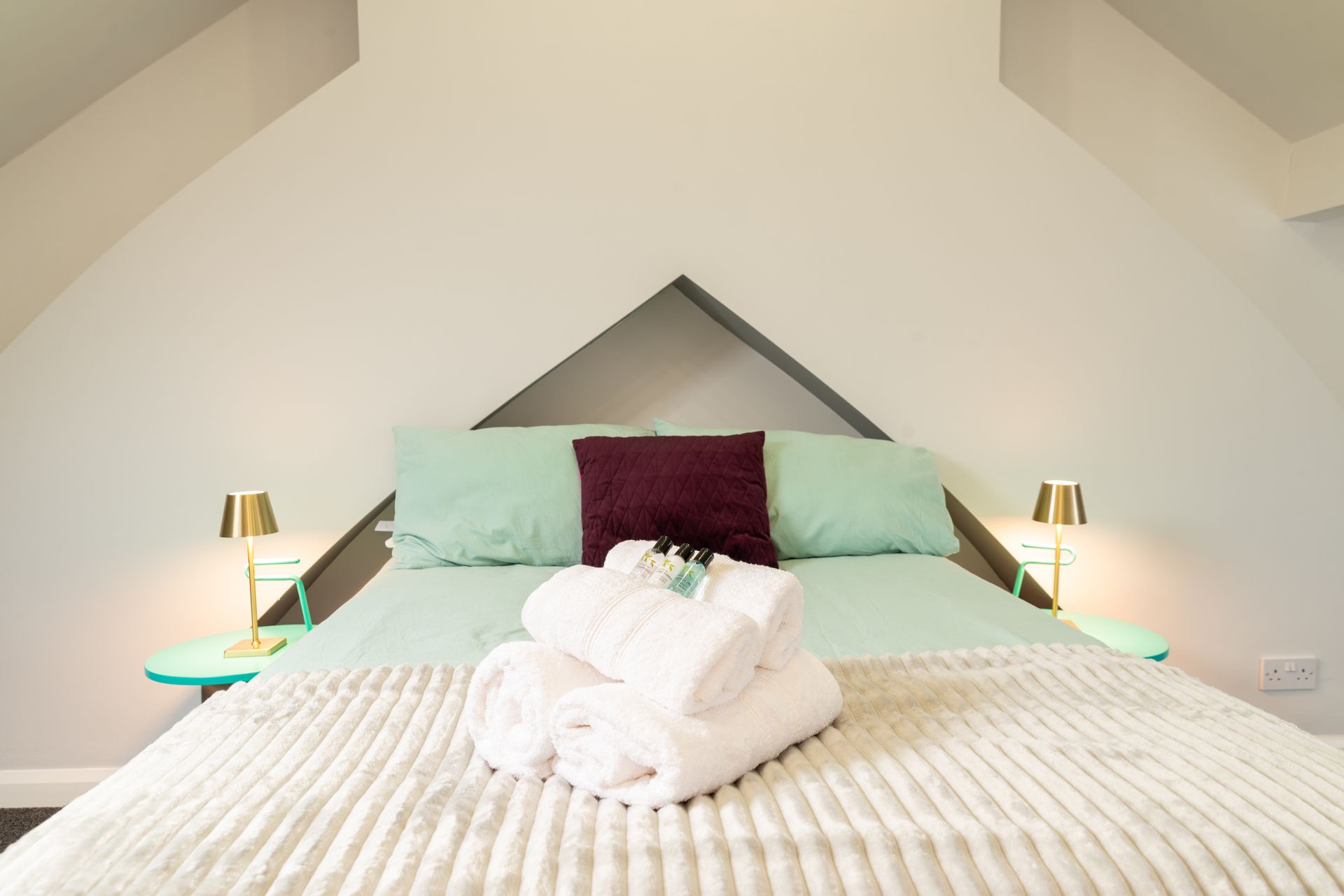 Cozy white bedroom with teal pillows, purple accent cushion, and folded towels on the bed