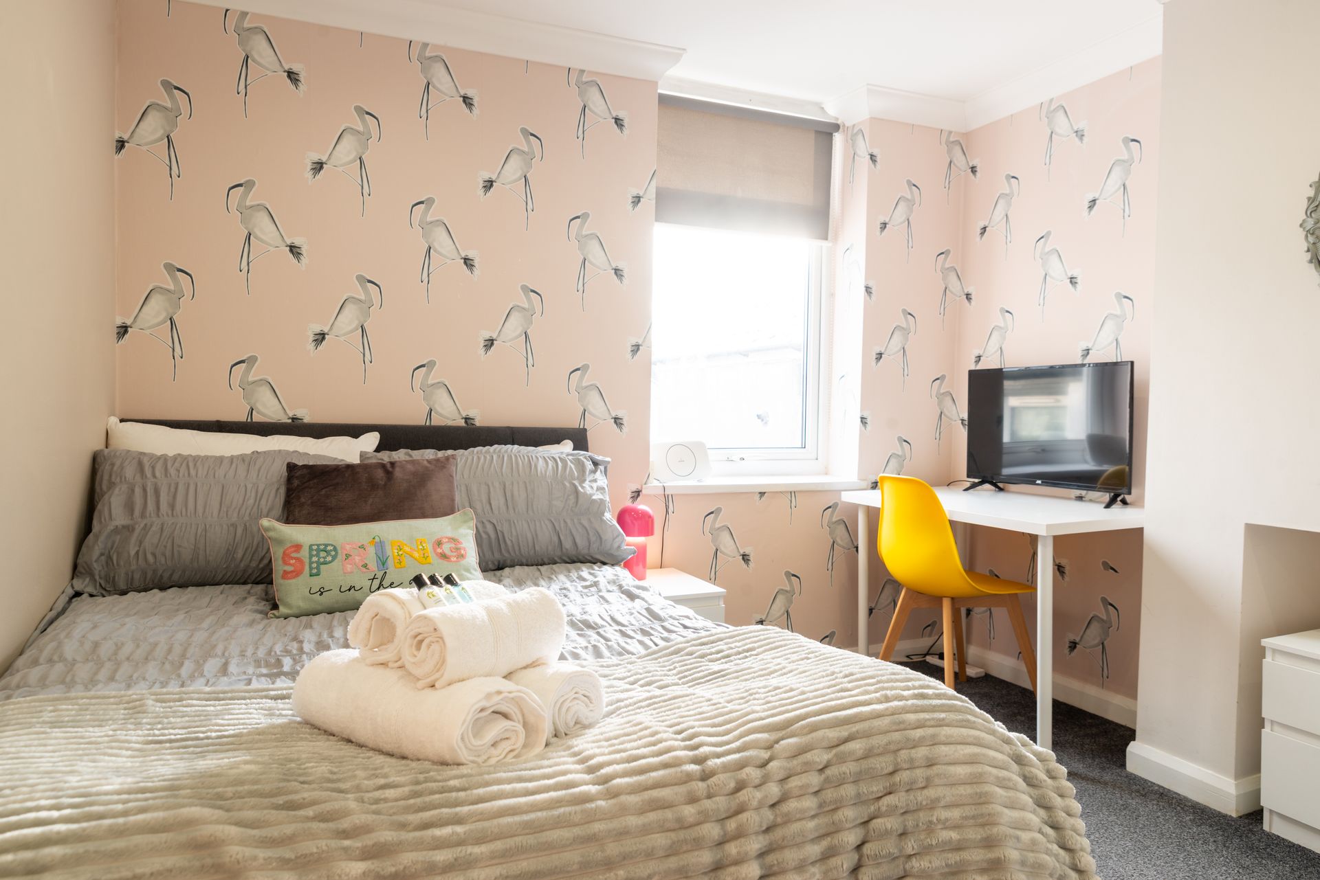 Bright bedroom with floral wallpaper, made bed, white desk with monitor, and yellow chair by the window.