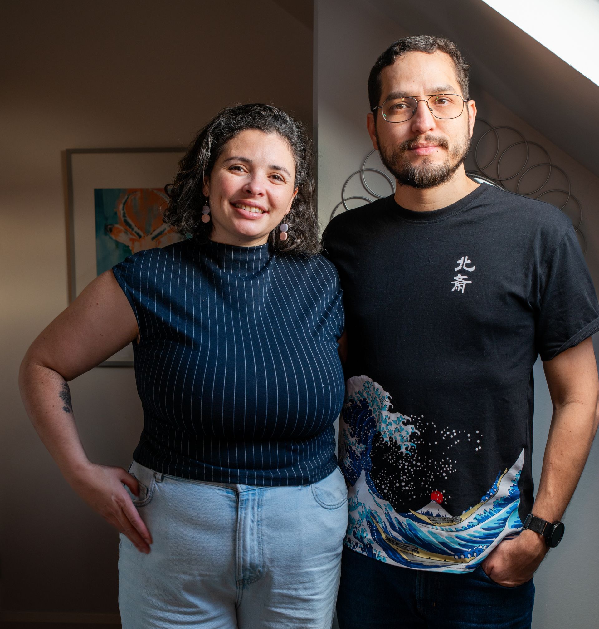 Two people standing indoors, smiling at the camera; one wears a black shirt and the other a sleeveless navy top.
