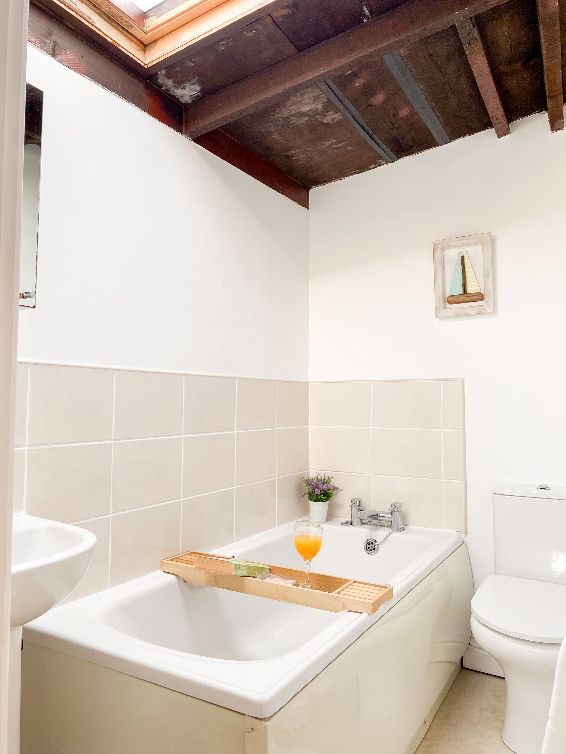 Small bathroom with a white bathtub, toilet, tiled walls, and wooden ceiling beams.