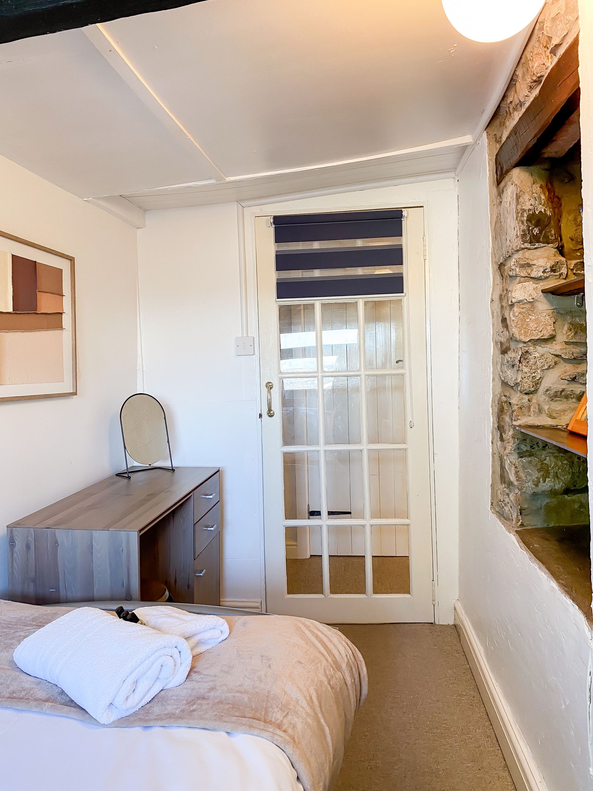 Small bedroom with bed, wooden dresser, mirror, and door with striped blind.